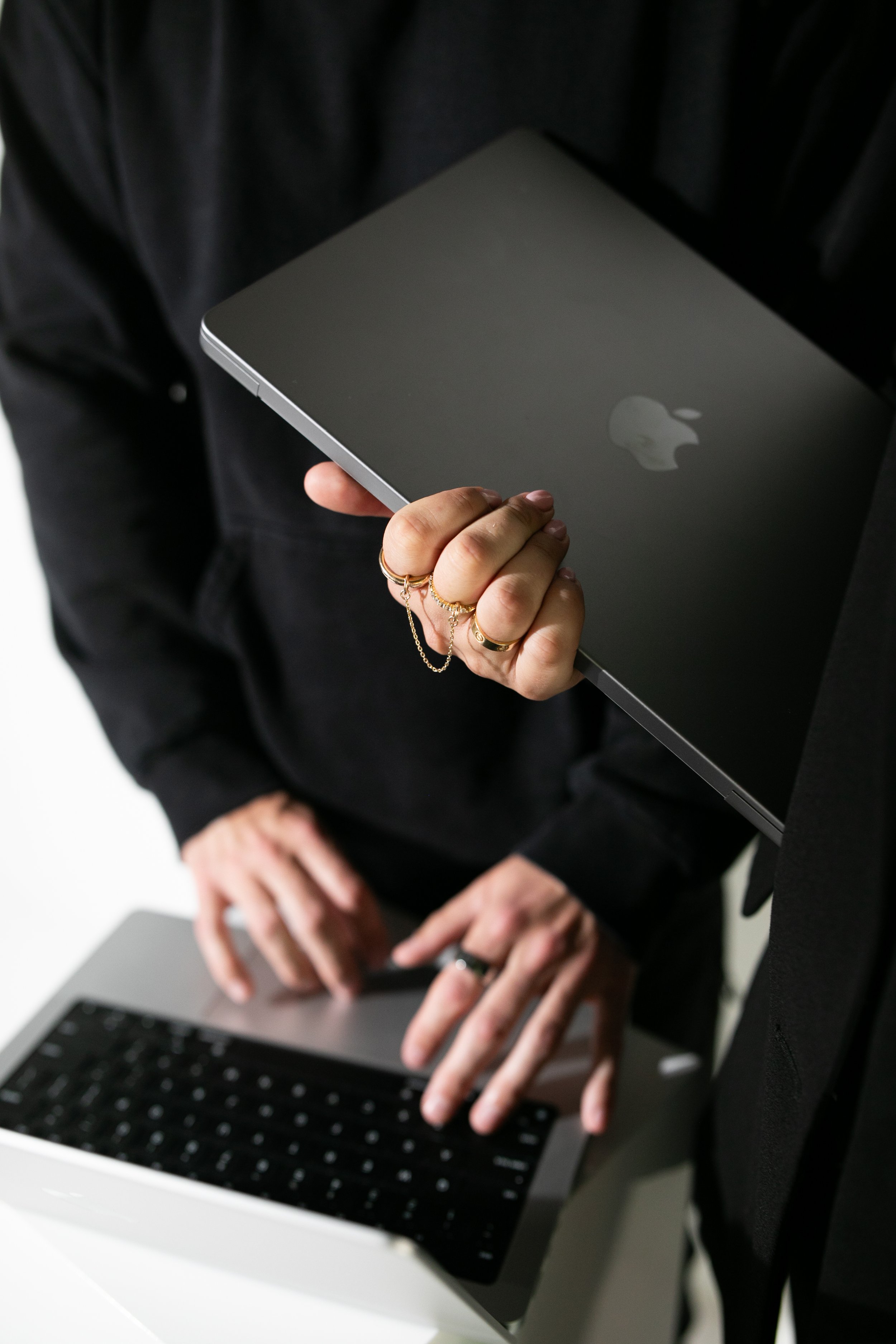 A person holding a closed MacBook with the Apple logo on top, standing near a laptop with their other hand in the background.