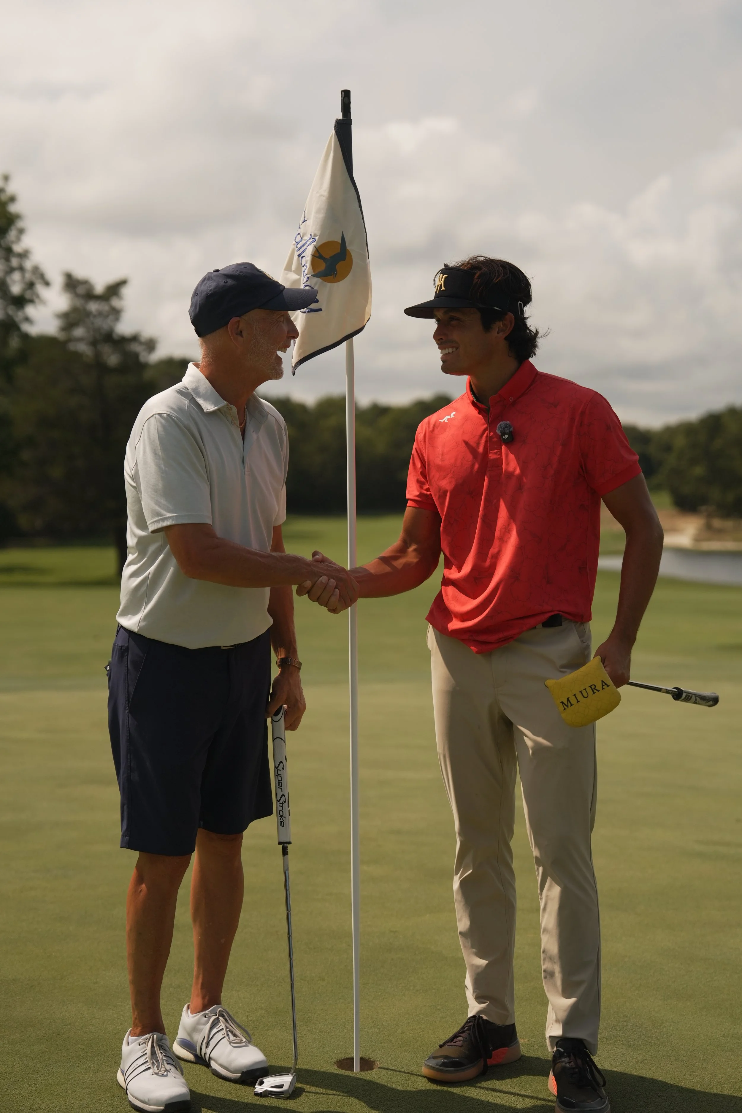 Two men shaking hands on a golf course, one dressed in a white polo and shorts, holding a golf club, and the other in a red polo and khakis, both wearing visors. They are near a golf hole with a flag.