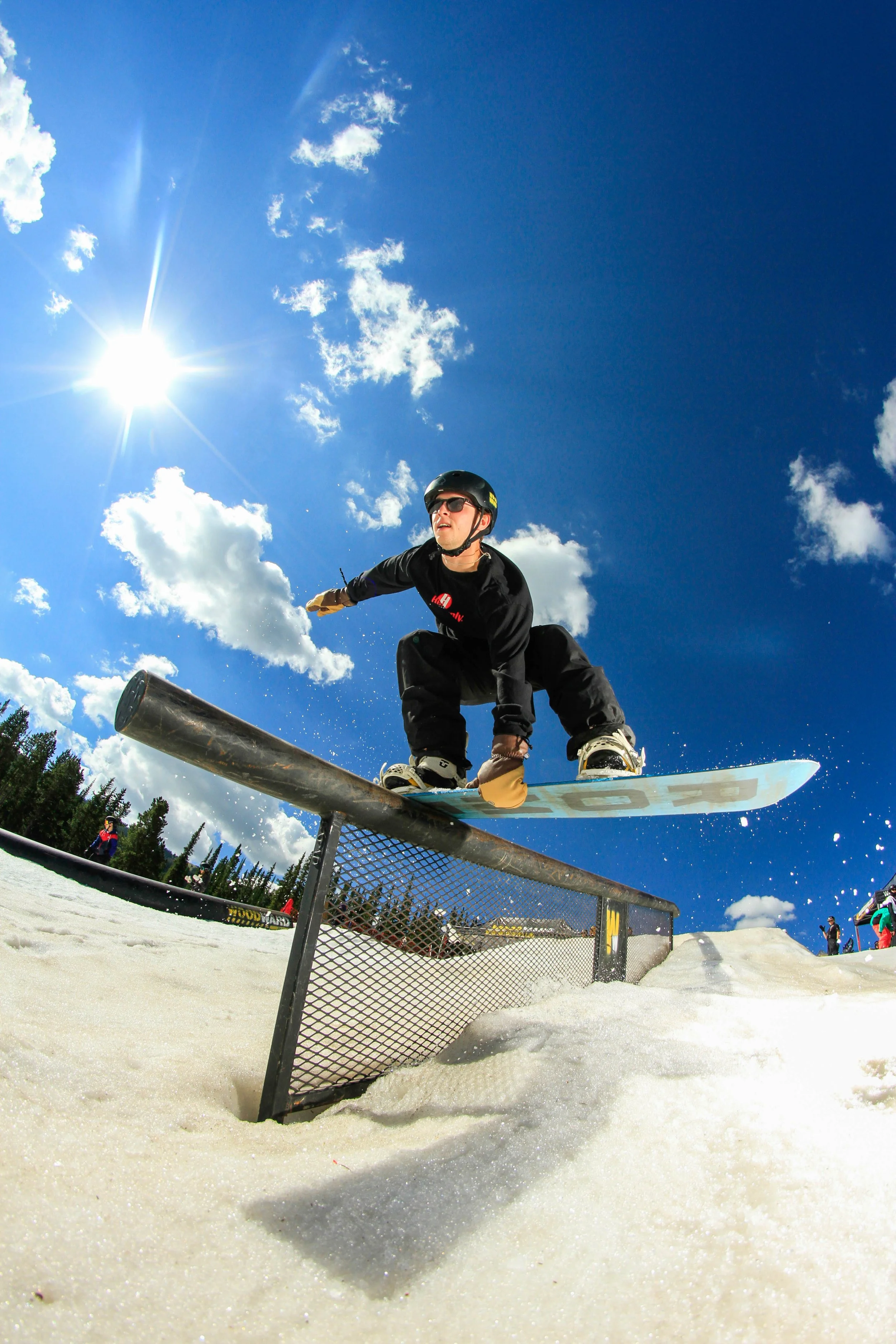 Snowboarder sliding across a metal rail on a sunny spring day, wearing a helmet and gloves, with bright blue sky and scattered clouds overhead.