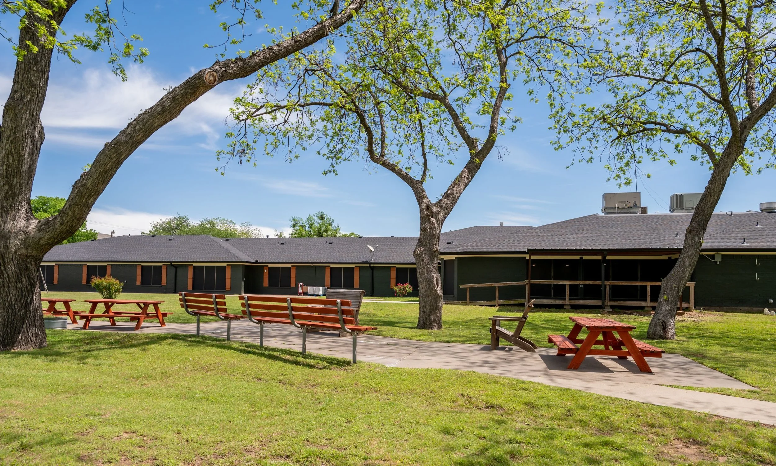 A park with green grass, trees, and a paved walking path. There are red picnic tables and benches, and a single-story building with a dark roof in the background. The sky is blue with some clouds.