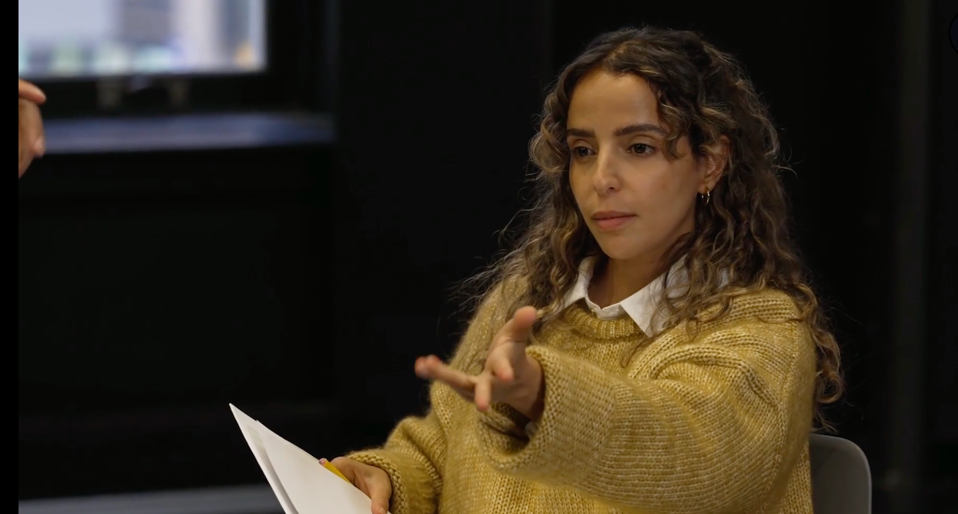 A woman with curly brown hair, wearing a white shirt and a mustard sweater, sitting in a dark room, gesturing with her hand, and holding papers.