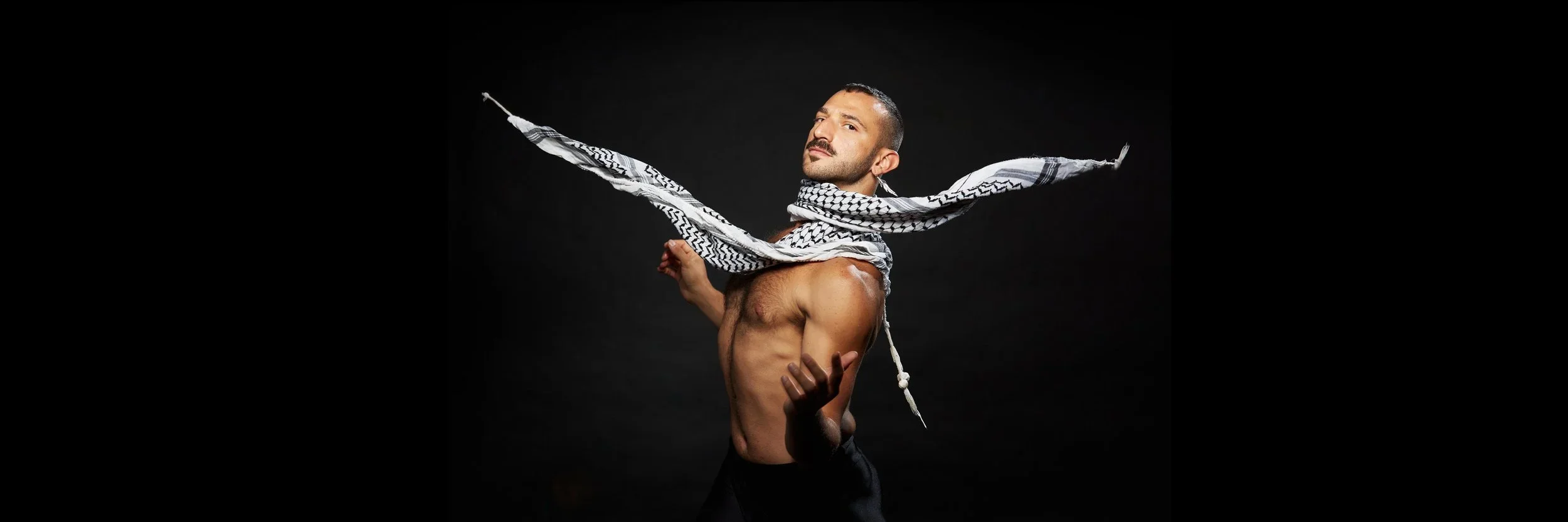 A shirtless man with a patterned scarf around his neck, posing against a black background.