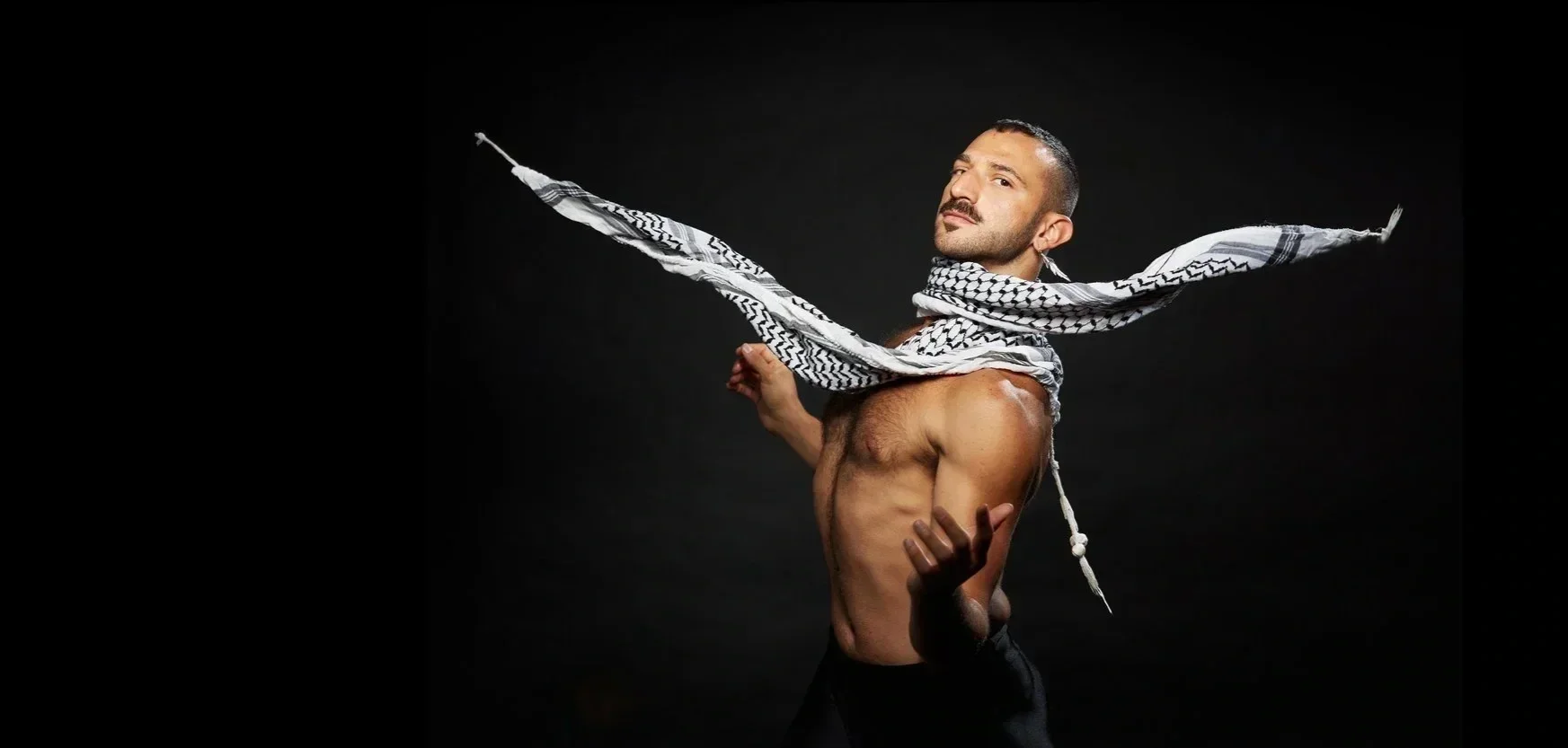 A shirtless man with a patterned scarf around his neck, posing against a black background.