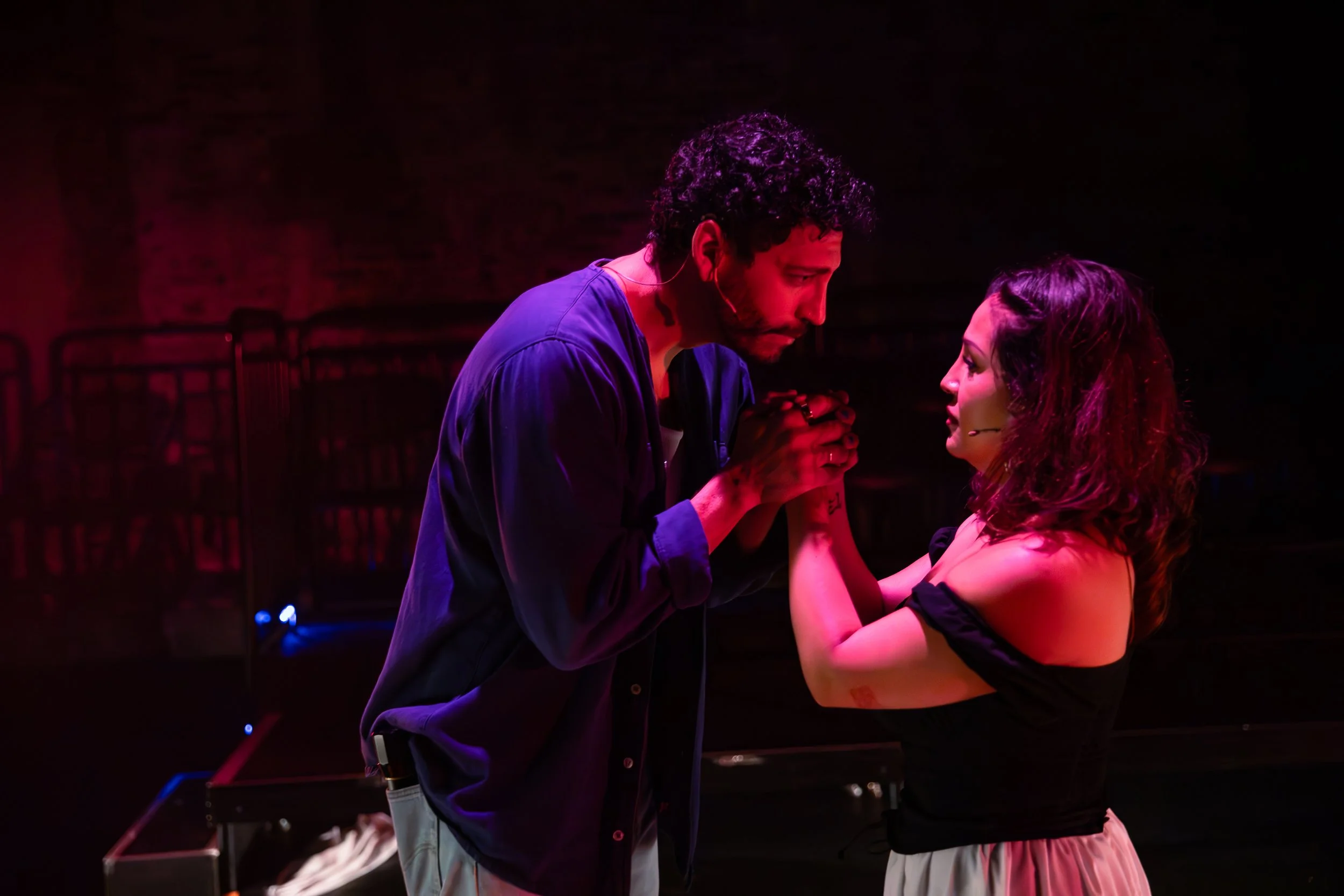A man with curly hair, wearing a microphone headset, is holding hands with a woman with dark hair, wearing a black dress. They are looking into each other's eyes in a dark stage setting with red and purple lighting.