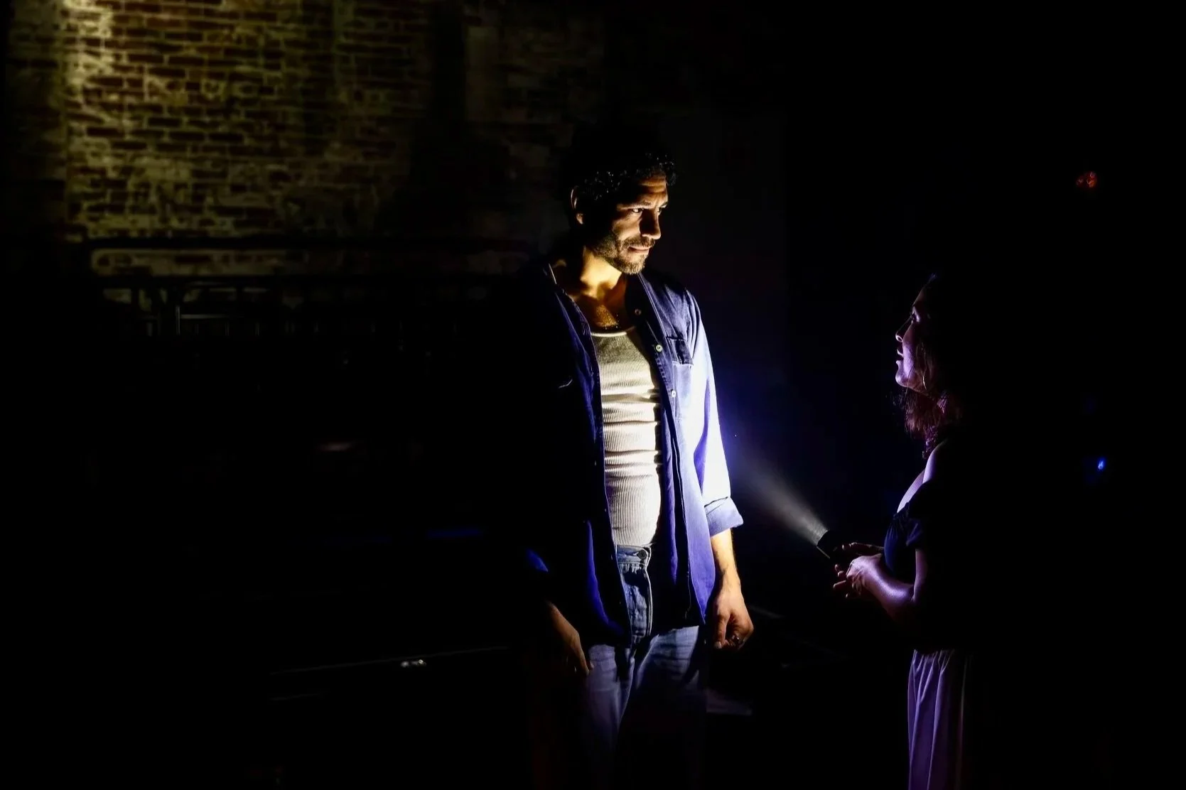 A man with curly hair, wearing a microphone headset, is holding hands with a woman with dark hair, wearing a black dress. They are looking into each other's eyes in a dark stage setting with red and purple lighting.