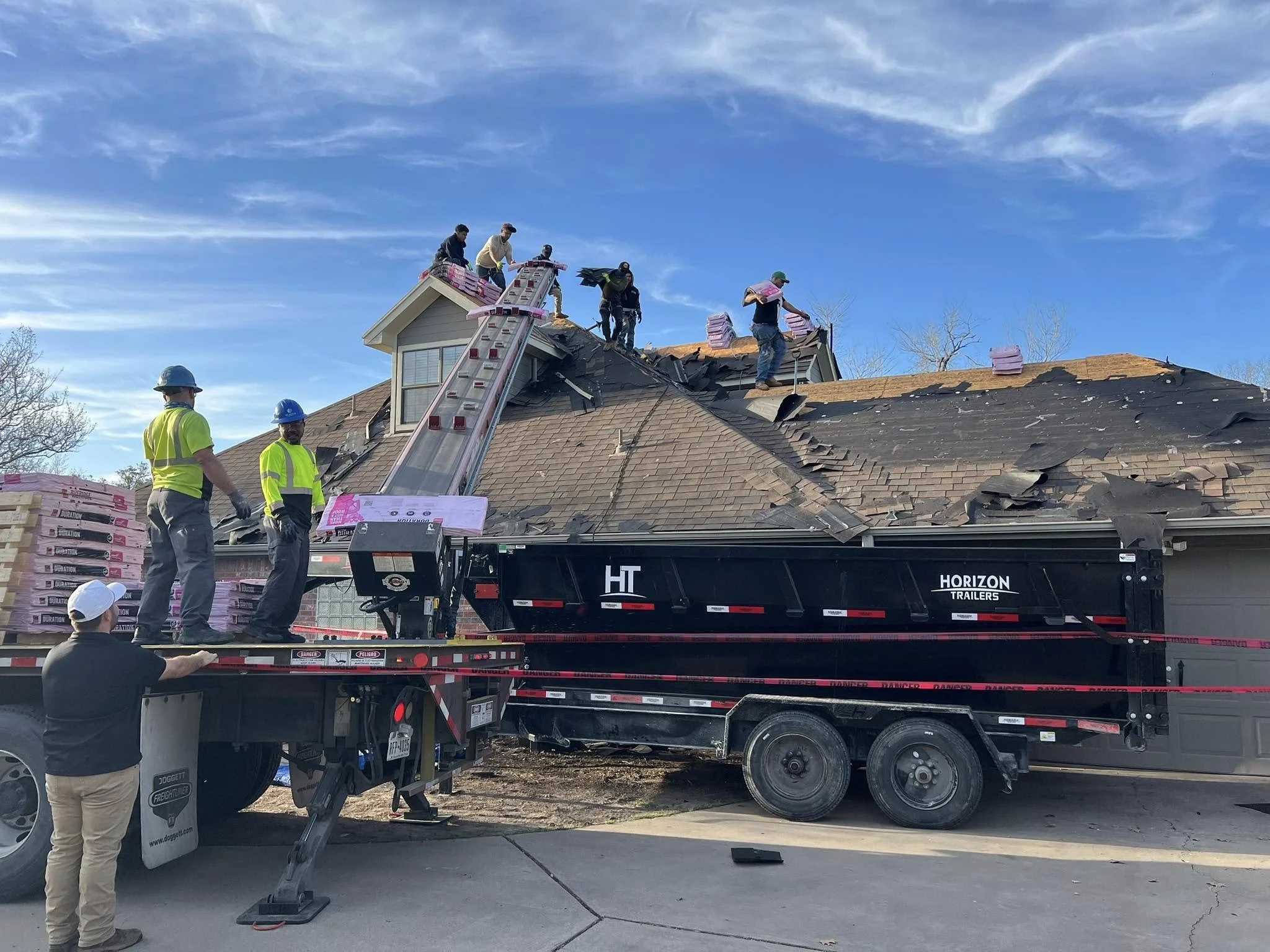 Workers on a ladder repairing a damaged roof, with some standing on the ground observing, and a flatbed truck nearby unloading roofing materials on a sunny day.