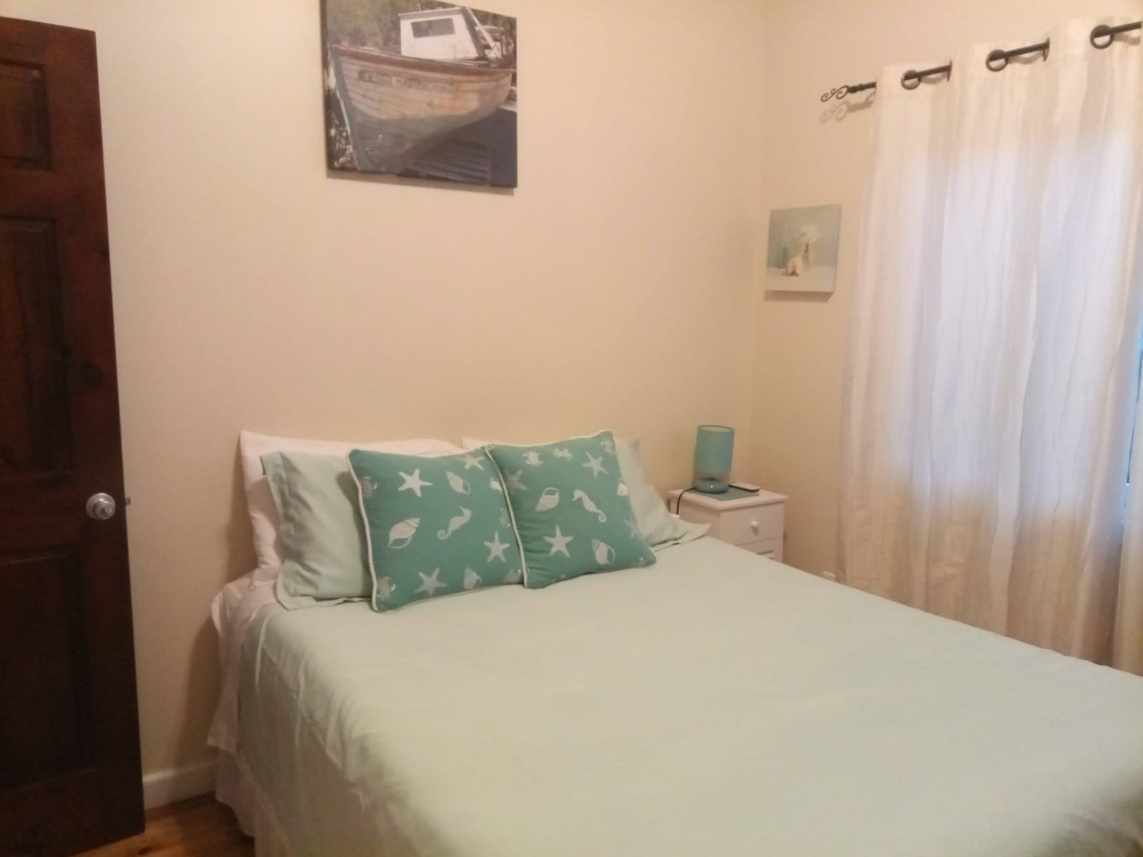 A bedroom with a bed, two sea-themed pillows, a small white nightstand with a teal lamp, cream curtains, and a wall art of a dog, with a wooden door and a picture of a boat on the wall.