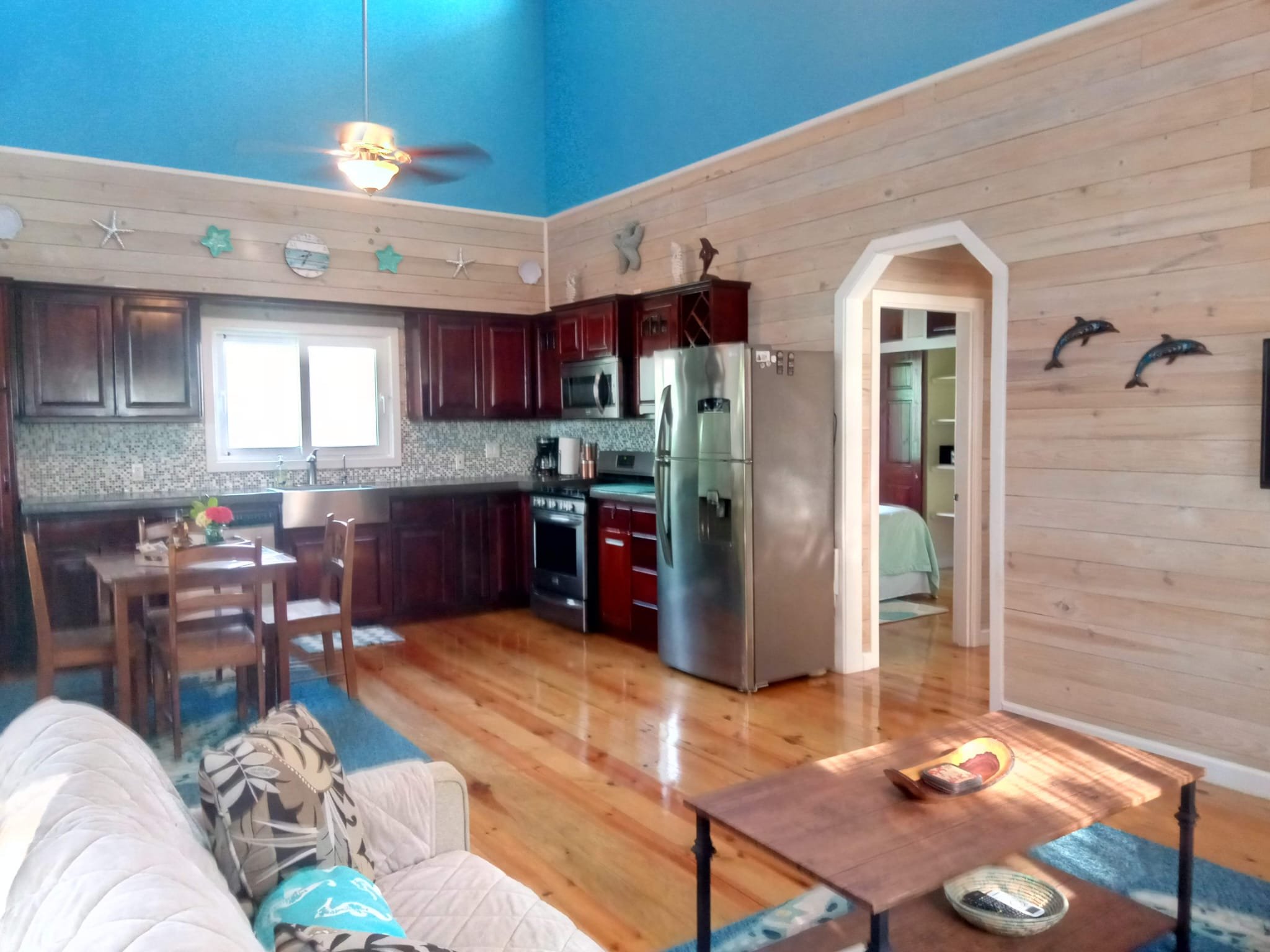 Open concept kitchen and living room with wood floors, dark wood cabinets, stainless steel appliances, and a blue ceiling. The living area has a beige sofa with patterned pillows, a wooden coffee table, and a wall-mounted TV. The kitchen has a small