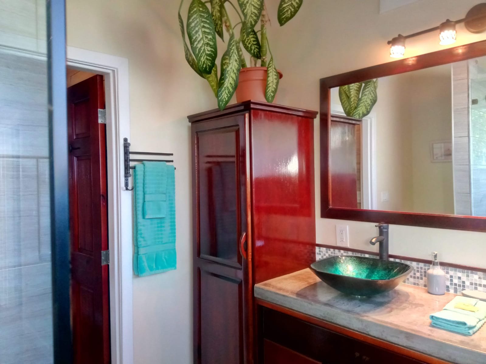 Bathroom interior with a large mirror, a green glass sink, a soap dispenser, a towel, a tall wooden cabinet, a plant on top of the cabinet, and a towel rack with a blue towel hanging.