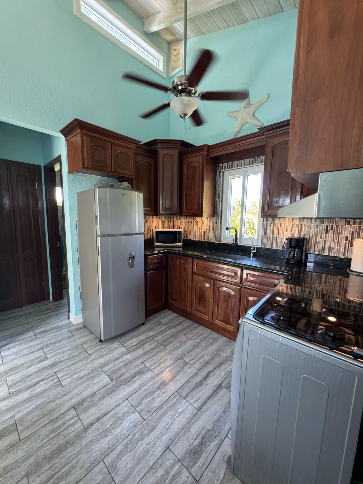 Kitchen with wooden cabinets, black countertop, white refrigerator, stove, microwave, coffee maker, window, ceiling fan, starfish decoration, wood-look tile floor, and high ceiling.
