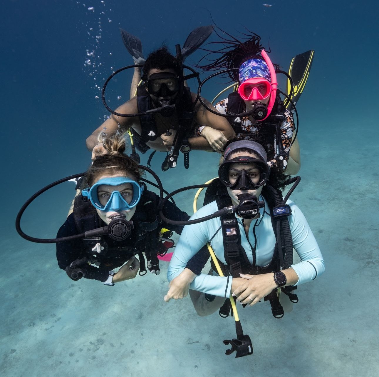 Four scuba divers underwater wearing masks and wetsuits, showing close-up of their faces and equipment.