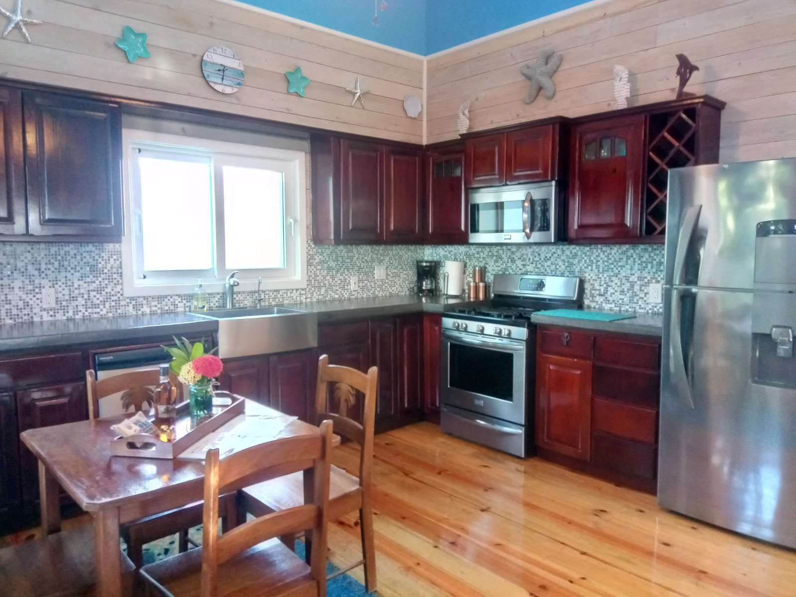 Kitchen with wooden cabinets, stainless steel appliances, a window above the sink, a wooden dining table with chairs, and decorative nautical items on the wall.