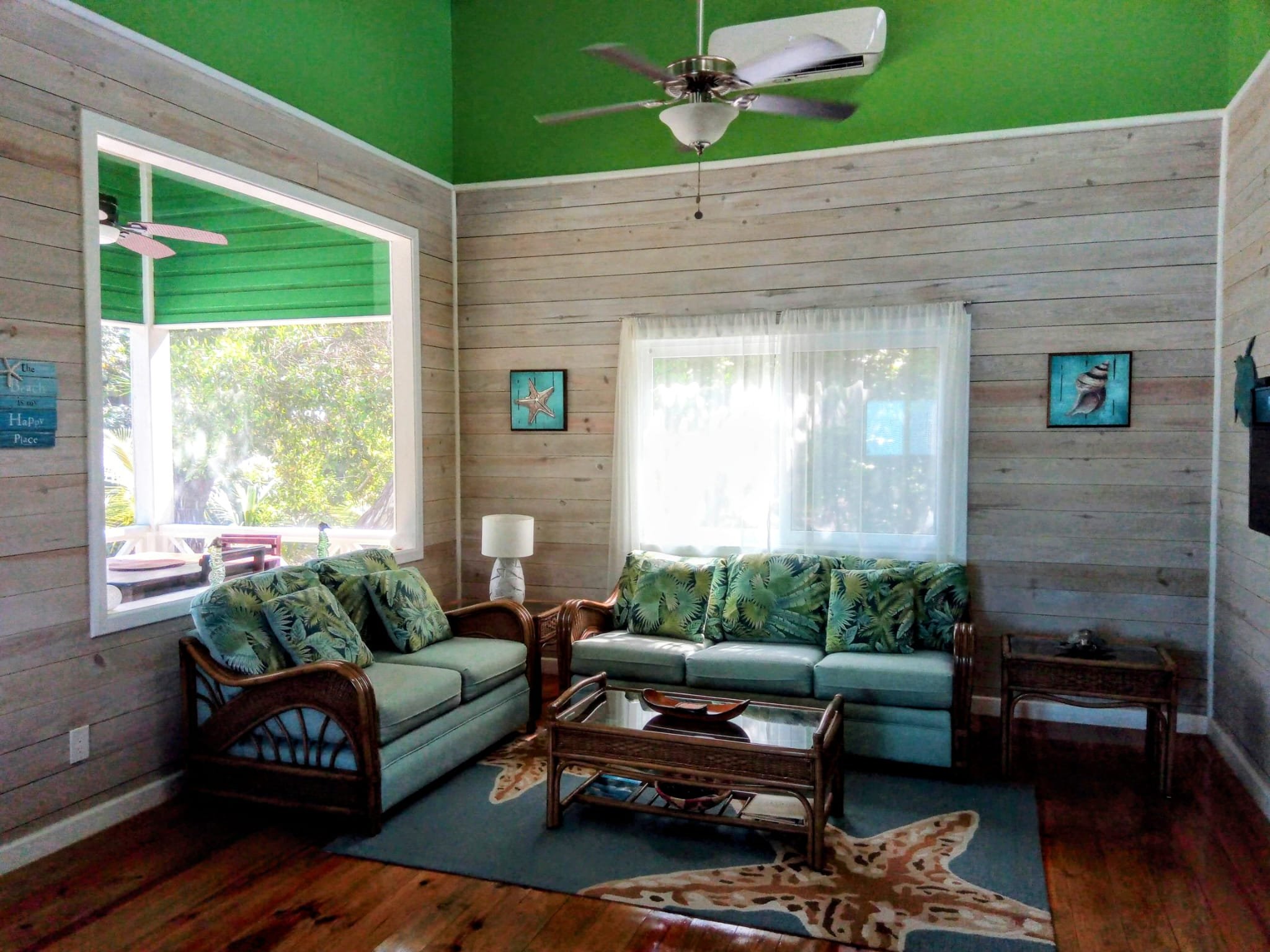 Living room with two rattan sofas with tropical leaf cushions, wooden coffee table, side table, a white floor lamp, and ocean-themed artwork on the wood-paneled walls. Large windows reveal greenery outside, and the ceiling is painted green with a cei