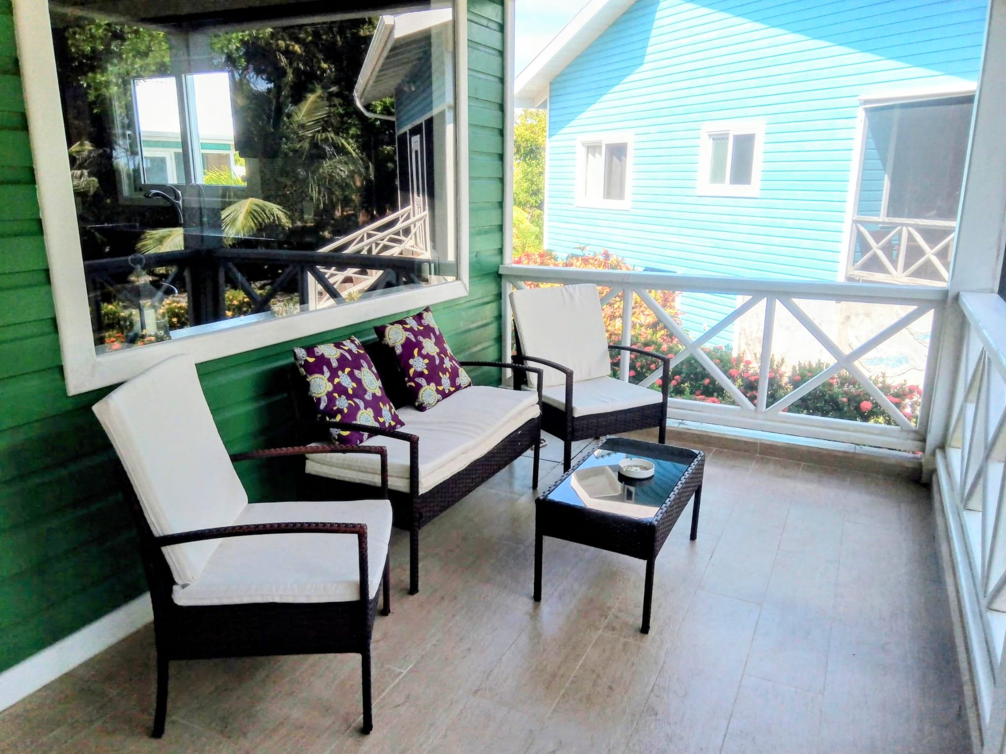Enclosed porch with white wicker chairs, a black wicker loveseat, and purple cushions with seashell patterns. Large windows show reflections of trees outside, and there are colorful houses visible beyond the porch railing.