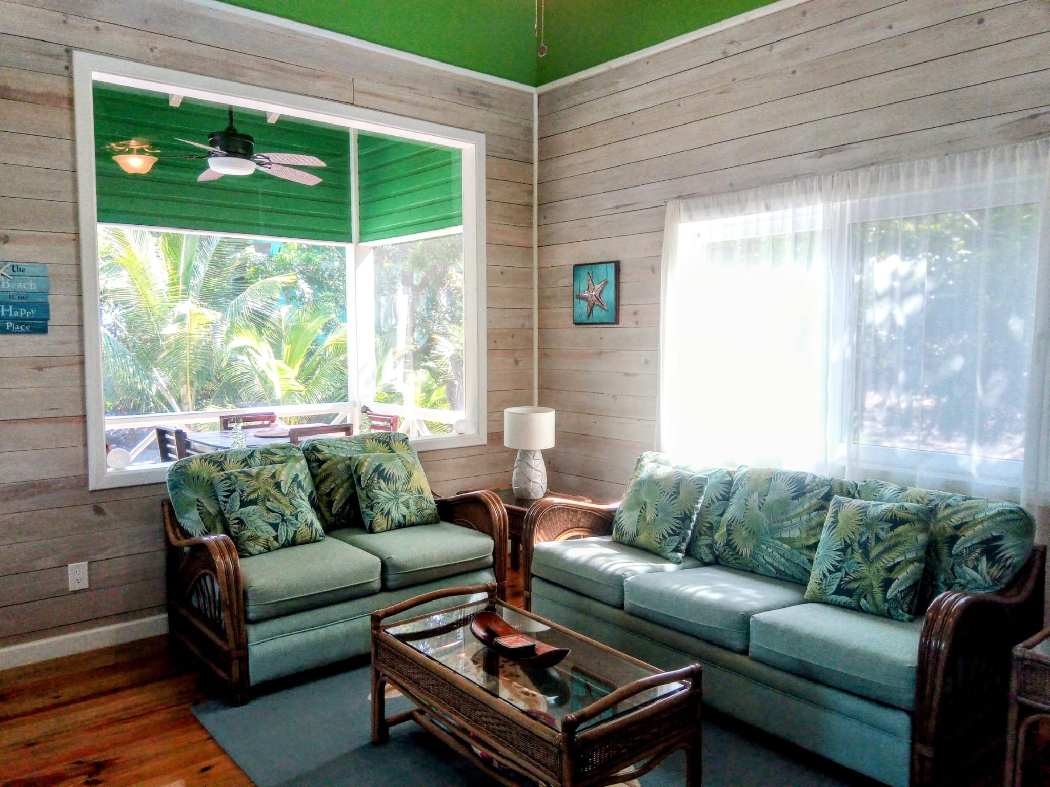 Living room with tropical decor, two light blue sofas with leafy patterned cushions, wooden coffee table, lamp, and large windows showing lush greenery outside.