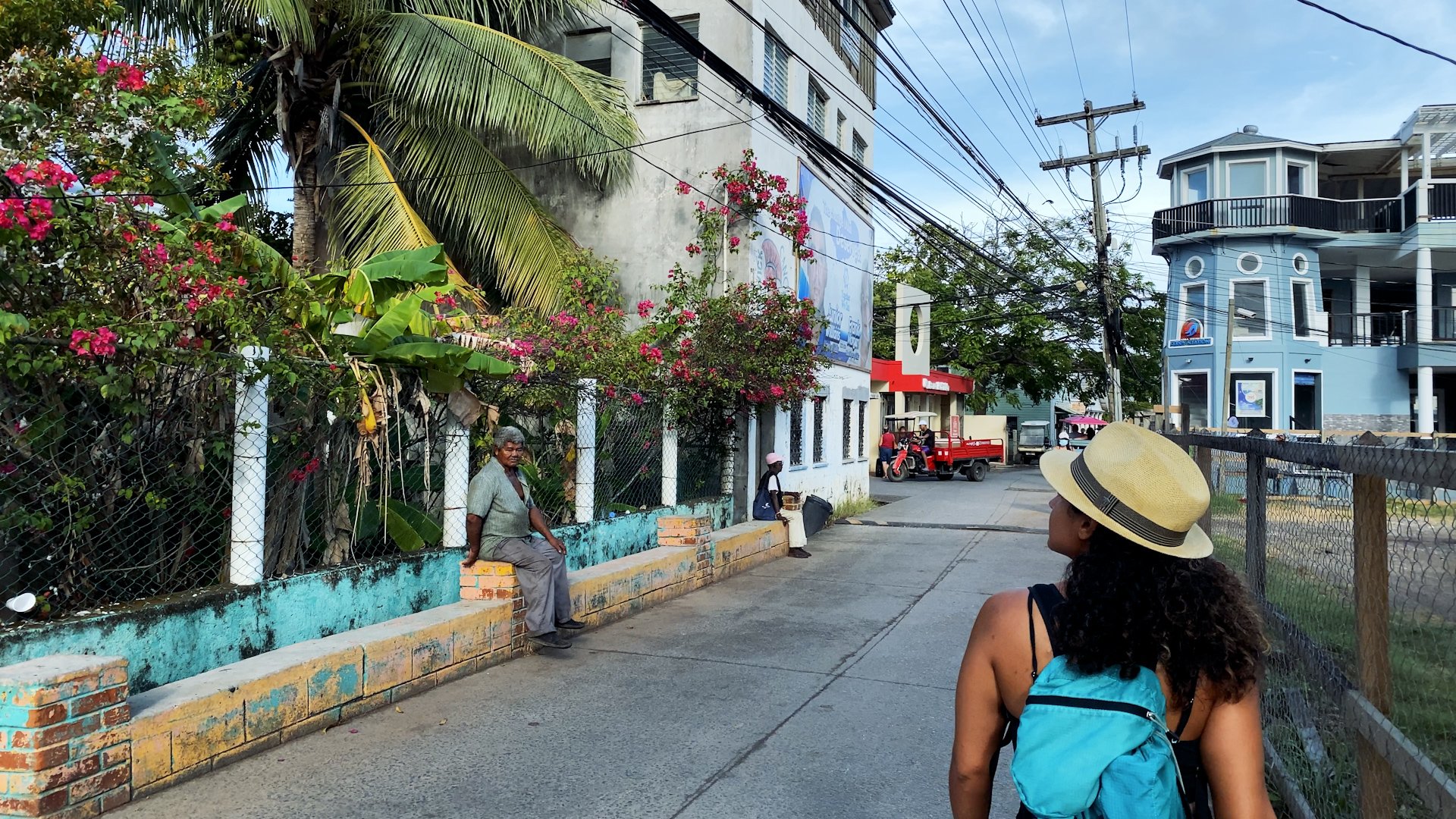A woman with curly hair, wearing a straw hat and a blue backpack, walks on a sidewalk past a white building with pink flowering bushes. An elderly man is sitting on a brick ledge near a wire fence with plants. A woman and a motorized tricycle are vis