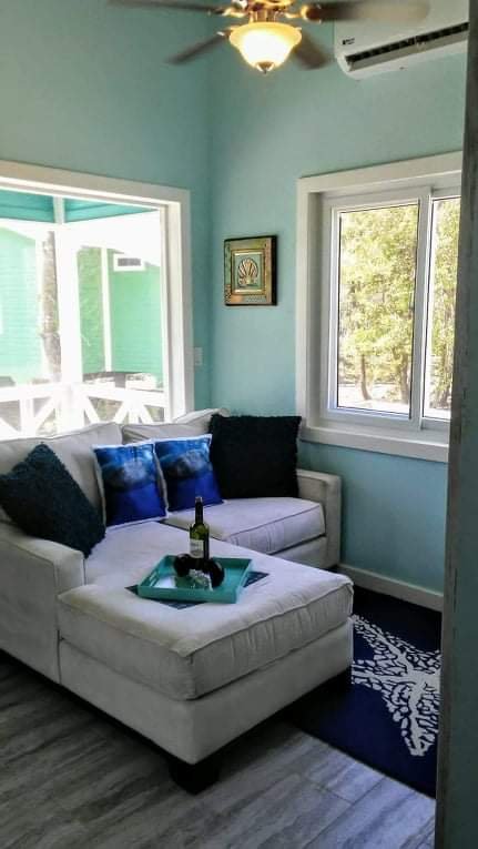 A cozy living room corner with a white sectional sofa adorned with black and blue throw pillows. A tray with a wine bottle and glasses is on the sofa's armrest. A window and sliding glass door let in natural light, with a view of green trees outside.