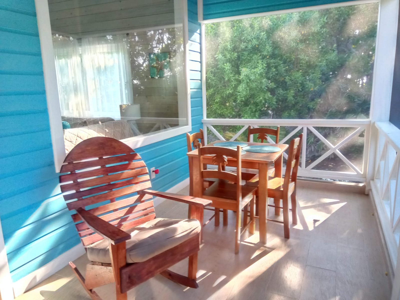 Sunlit screened porch with wooden table and chairs, and a wooden lounge chair, adjacent to a blue house with a large window, overlooking greenery.