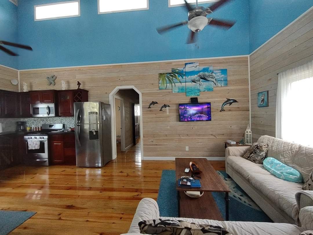 Living room with wooden floors, a beige couch with pillows, a wooden coffee table, a flat-screen TV on a wood-paneled wall with dolphin decorations, and a kitchen area with dark wood cabinets and stainless steel appliances, including a refrigerator.