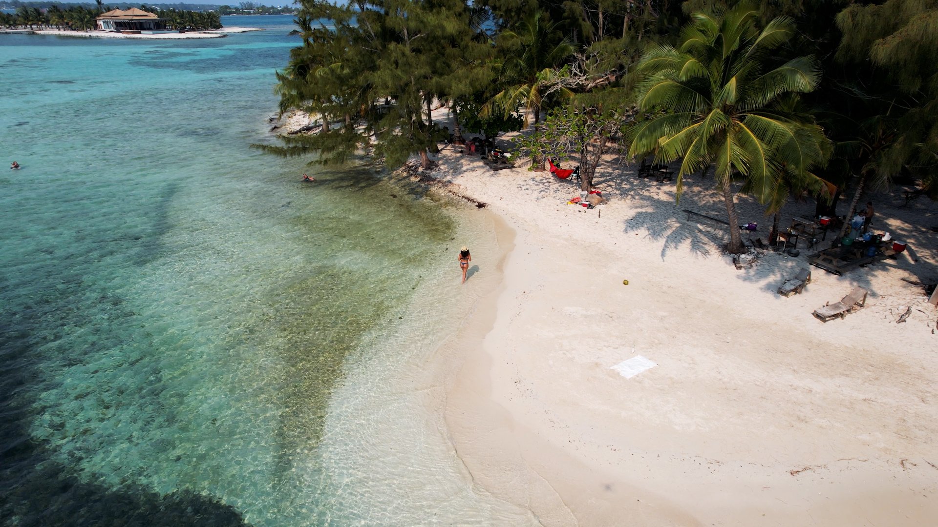 An aerial view of a tropical beach with white sand, turquoise water, and green palm trees. A woman in a hat walks along the shoreline, with some people swimming in the water and shadow of a building or trees cast on the sand.