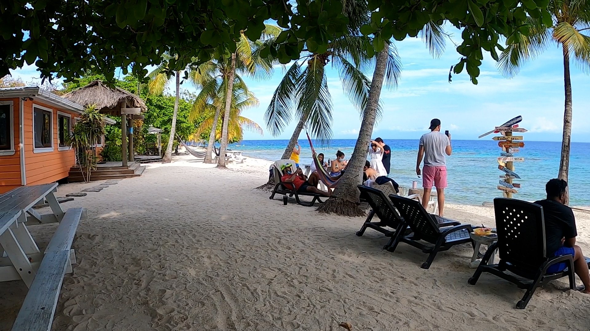 Beach scene with palm trees, a small orange hut, people relaxing on lounge chairs under the shade, some taking photos, a hammock, and a signpost near the water, with a blue ocean and sky in the background.