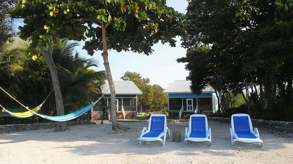 Three blue lounge chairs on a sandy beach in front of two small houses, with a hammock strung between trees and greenery surrounding the scene.
