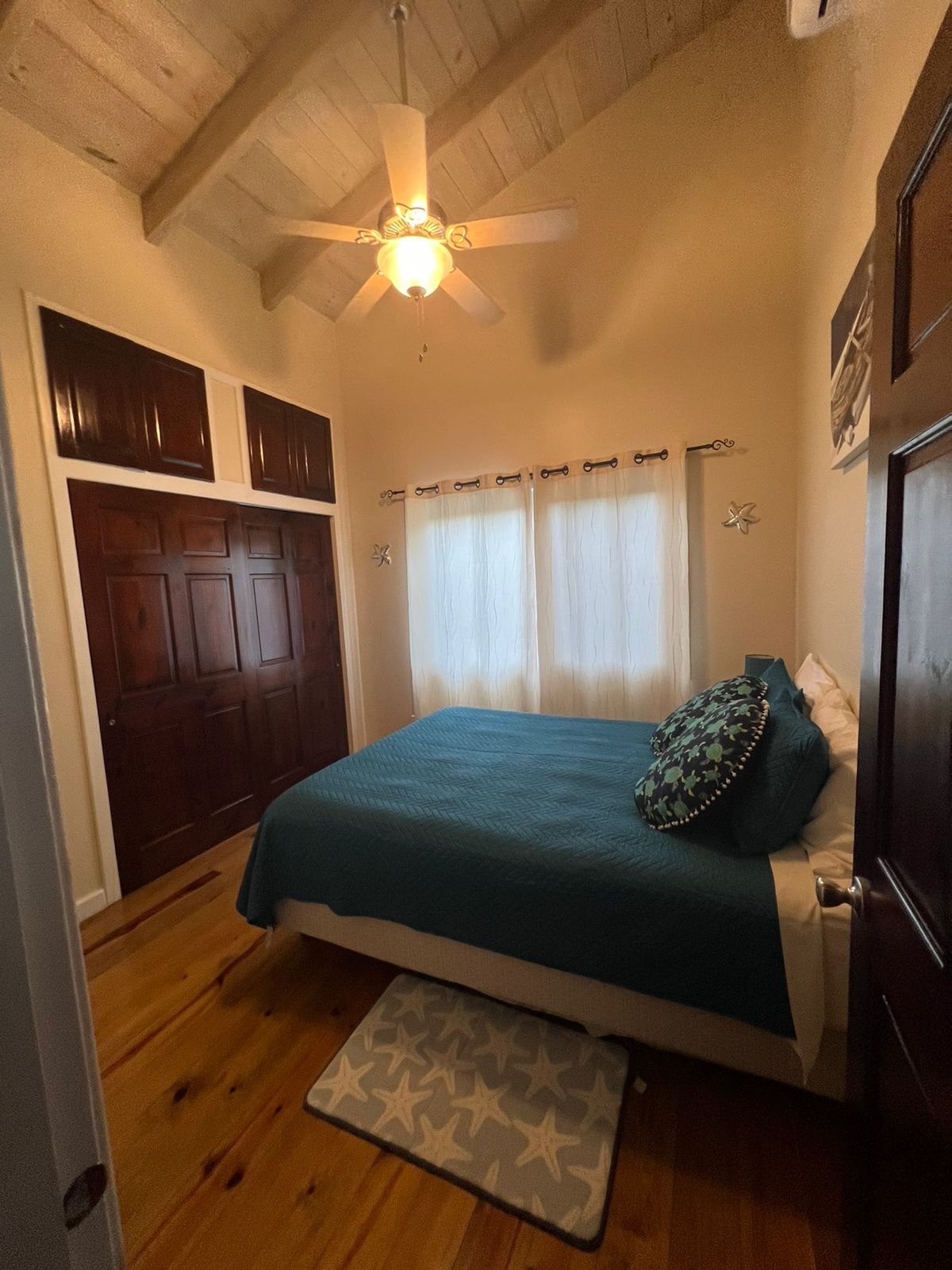 Cozy bedroom with a wooden vaulted ceiling, ceiling fan with light, cream walls, white curtains over a window, dark wood closet doors, a bed with blue bedding and coordinating pillows, and a cream starfish patterned rug on a wood floor.