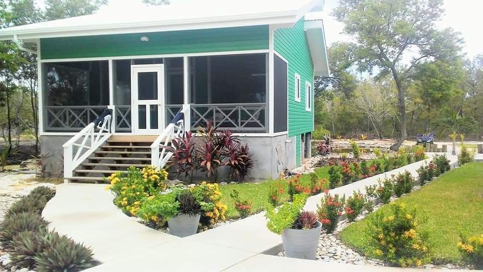 A small house with green siding, a screened porch, and white stairs leading up to it. The front yard has neatly groomed grass, colorful flowering plants, and potted plants lining the walkway. Trees are visible in the background.