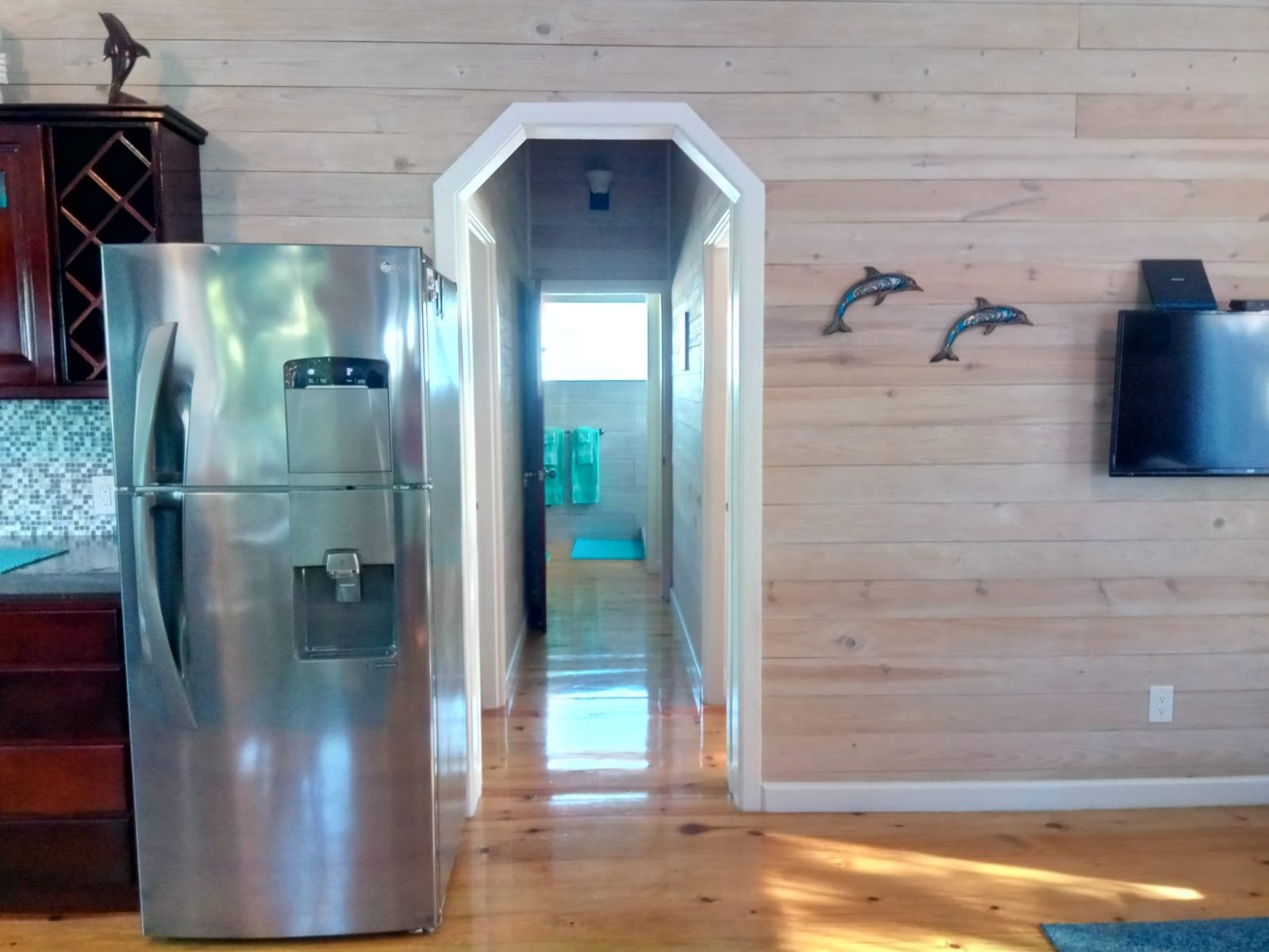 Interior of a wooden cabin with a stainless steel refrigerator, a wall-mounted flat-screen TV, and a hallway leading to a bathroom with a window and towels.