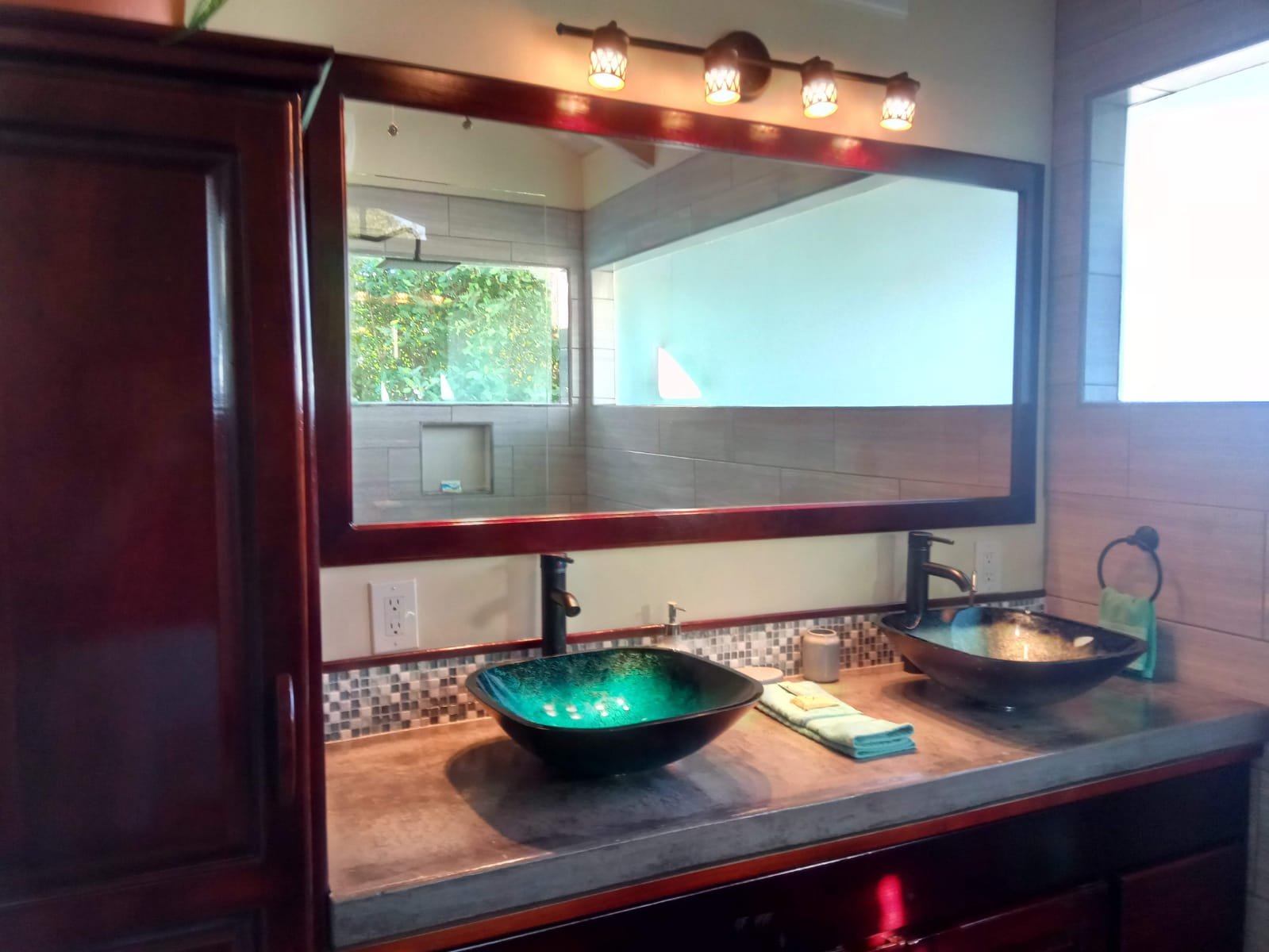Bathroom vanity with two black vessel sinks, a large mirror, two black faucets, and a countertop with folded towels and soap, wood cabinetry, tile backsplash, and a window providing natural light.