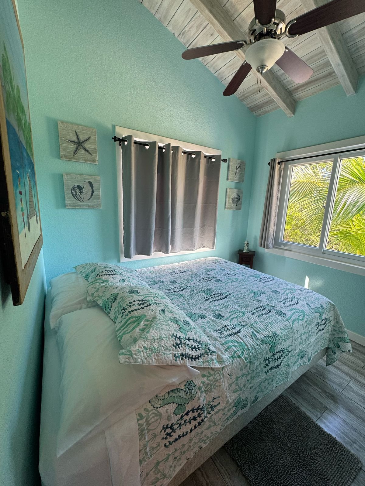 Coastal-themed bedroom with a bed covered in a sea-inspired quilt, light blue walls, a window with ocean view, gray curtains, a ceiling fan, and beach decor wall art.
