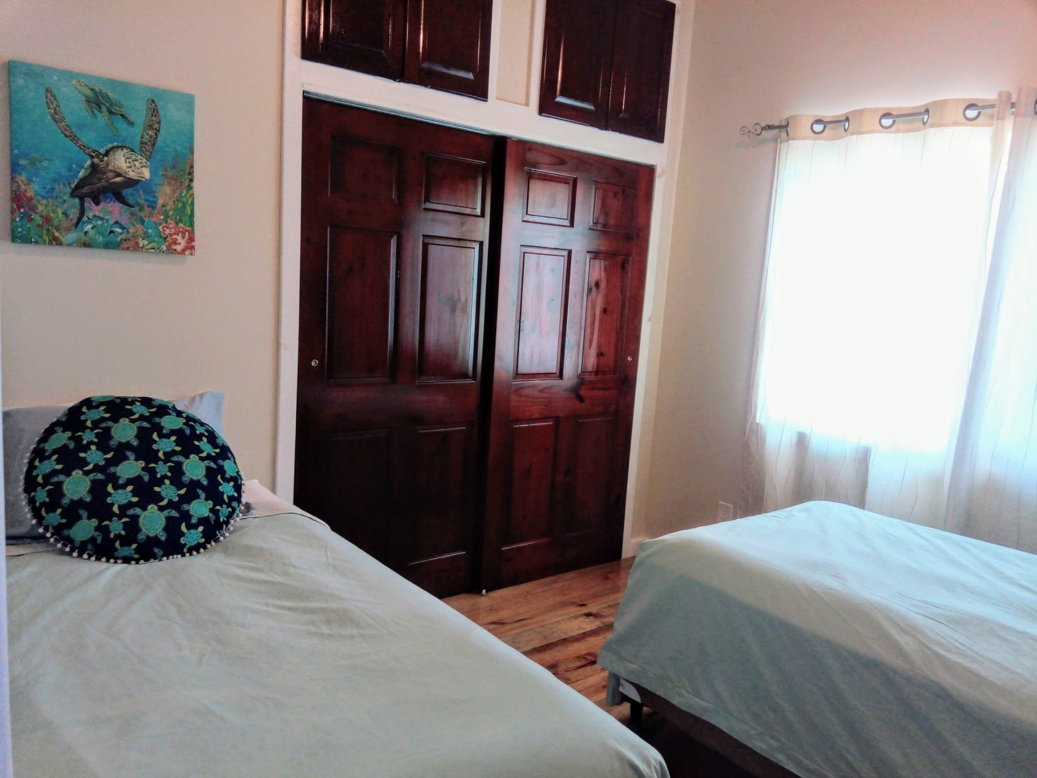 A bedroom with two beds, a window with sheer curtains, a painting of a turtle and other sea creatures, and a wooden closet with sliding doors.