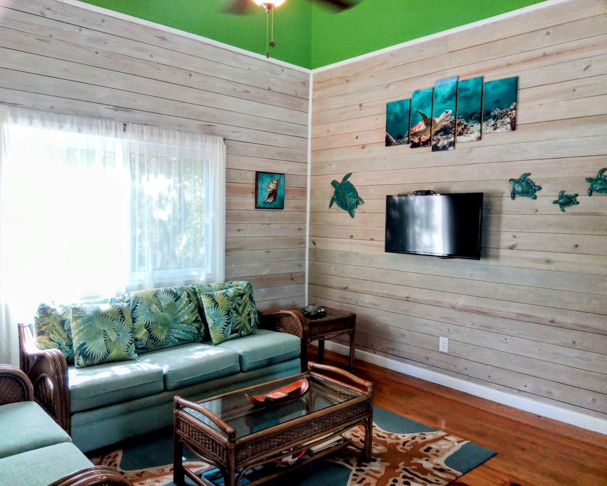 Living room with light wood panel walls, a window with white curtains, a green sofa with tropical leaf pillows, a glass-topped wicker coffee table, wall decorations of fish and sea turtles, a flat-screen TV, and a patterned area rug.