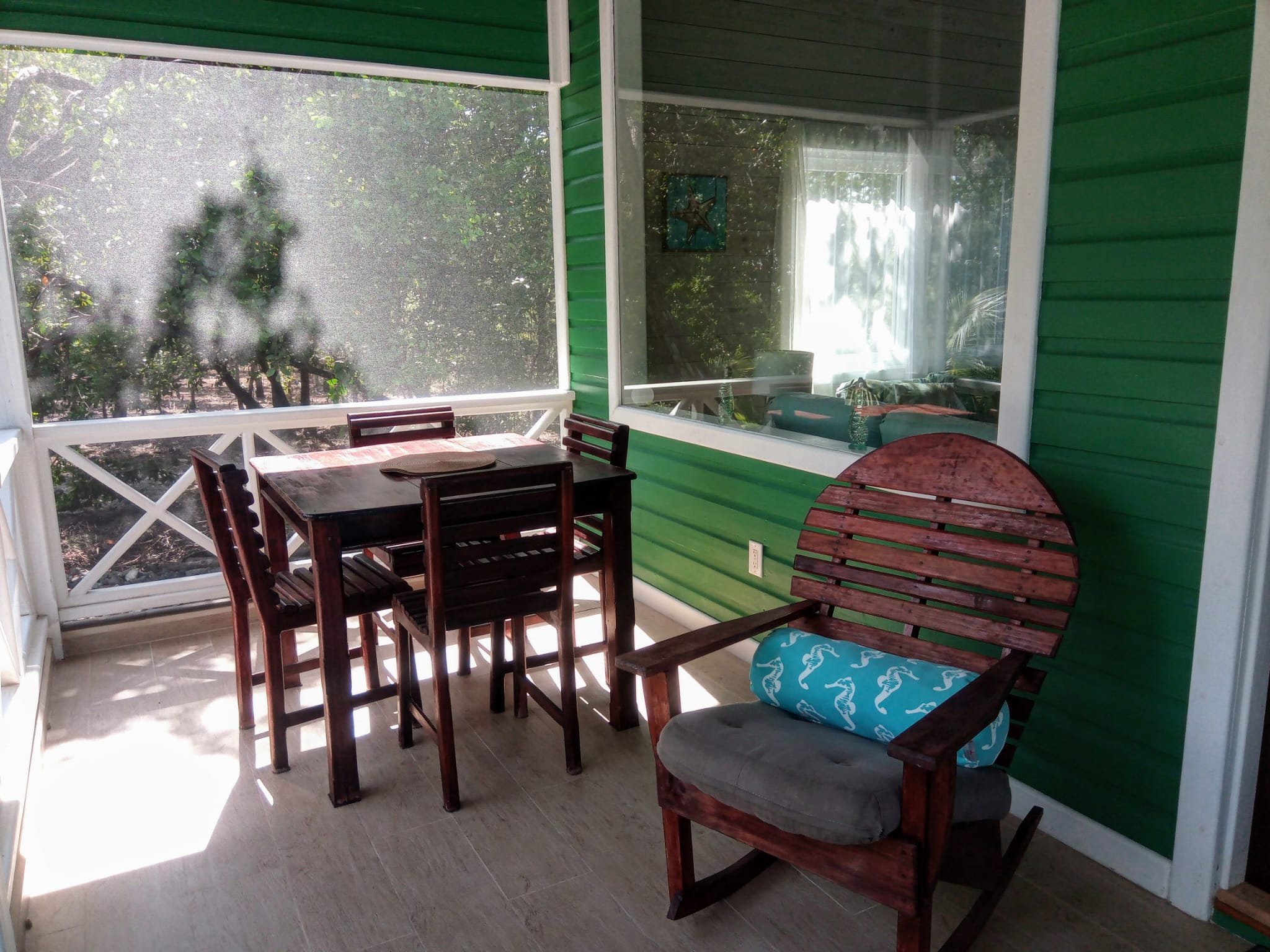 Porch with a wooden dining table and four chairs, green siding walls, a rocking chair with a seahorse pillow, and large windows overlooking trees and outdoor scenery.