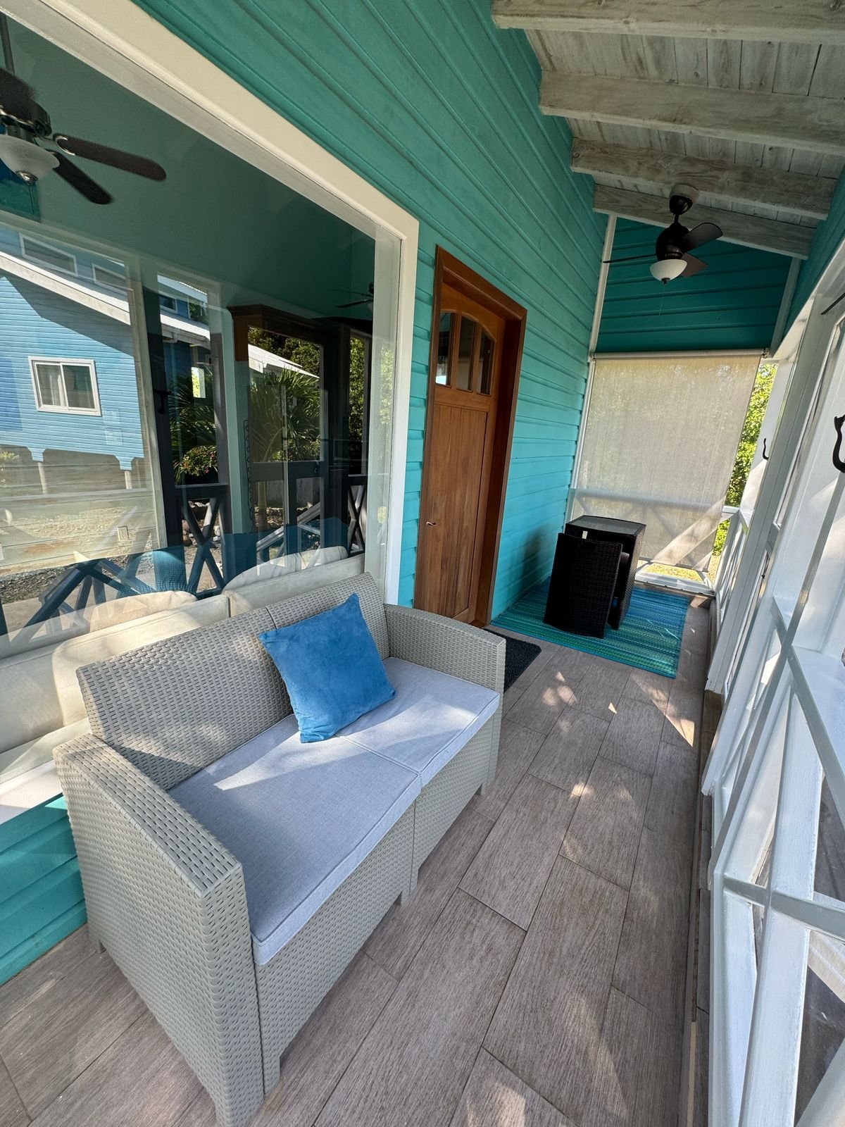 A small outdoor porch area with teal siding and wood flooring, featuring a gray wicker loveseat with a blue cushion, a glass door leading inside, and a ceiling fan. There's also a small black table and a striped rug at the far end.