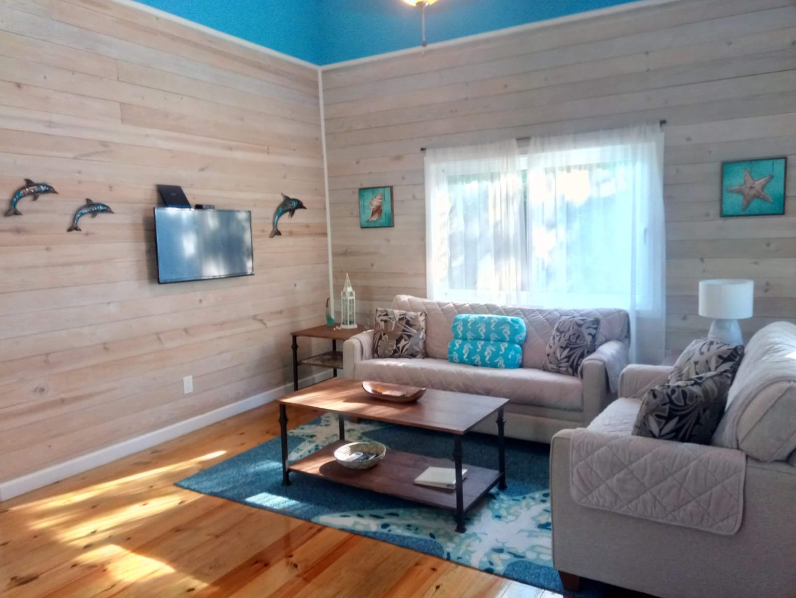 Cozy living room with light wood-paneled walls, a window with white curtains, two beige sofas with patterned pillows, a wooden coffee table, and sea-themed decorations including wall art and dolphins on the wall.