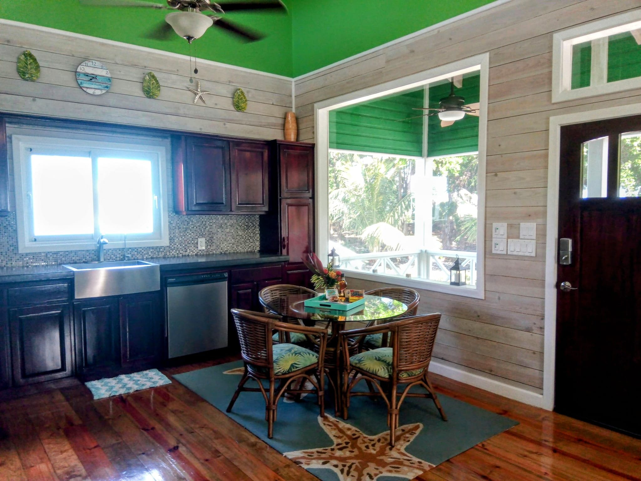 A kitchen with wooden cabinetry, a window above the sink, and a round dining table with four wicker chairs. The room has a green ceiling, wood-paneled walls, and a large window showing outdoor trees and greenery.