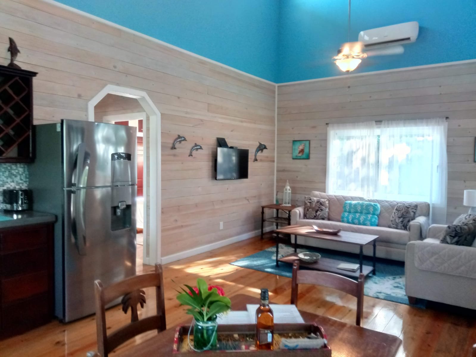 Living room with wood-paneled walls, a blue ceiling, a window with white curtains, a beige sofa with decorative pillows, a coffee table, a small side table with a lantern, a TV mounted on the wall, and a ceiling fan with a light fixture.
