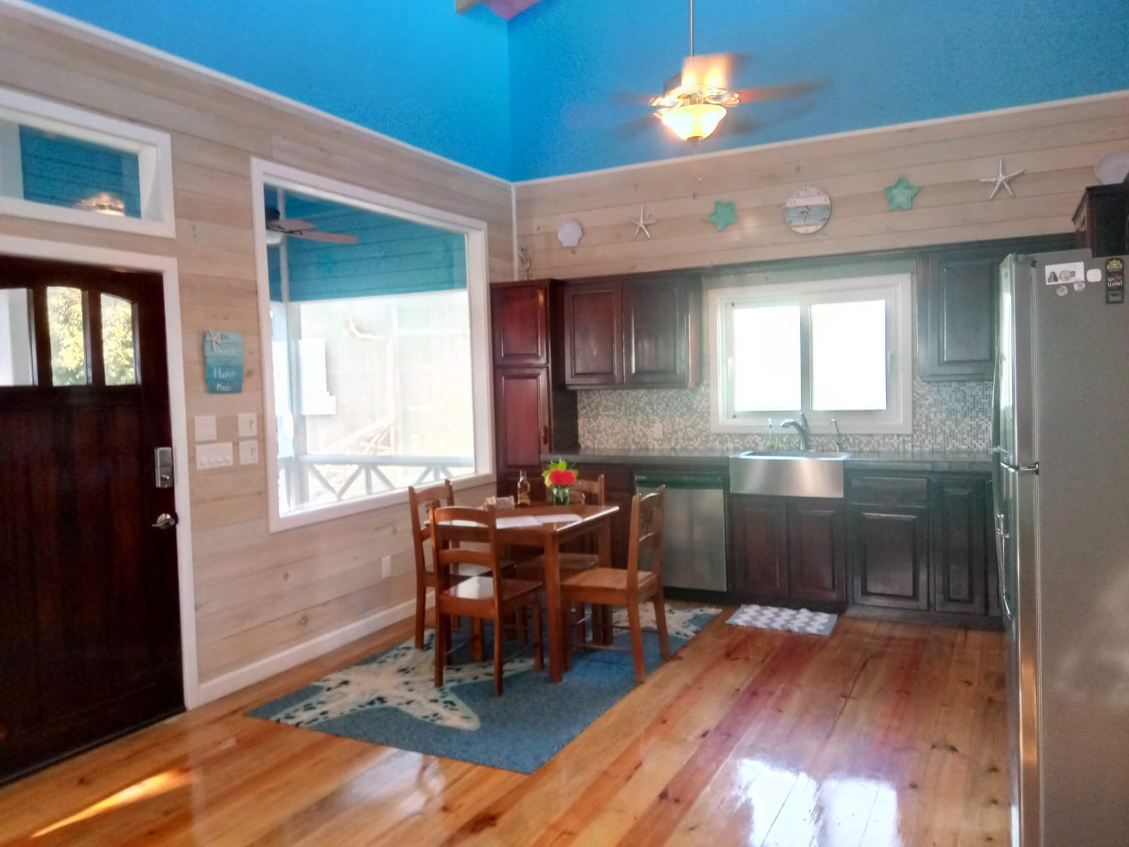 Cozy kitchen with wooden floors, dark wooden cabinets, a stainless steel refrigerator, and a small wooden dining table with four chairs. The walls are a mix of light wood paneling and bright blue paint, with beach-themed decor above the window.