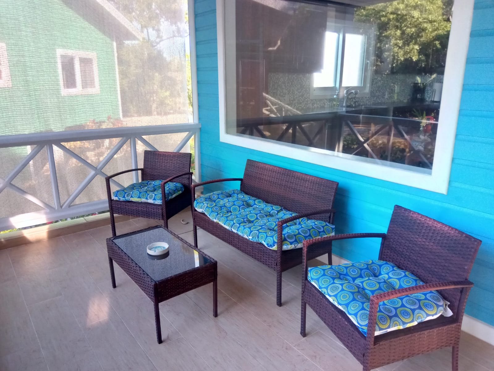Patio with wicker seating and a glass-topped table in front of a blue house with large window, overlooking a green house and trees outside.
