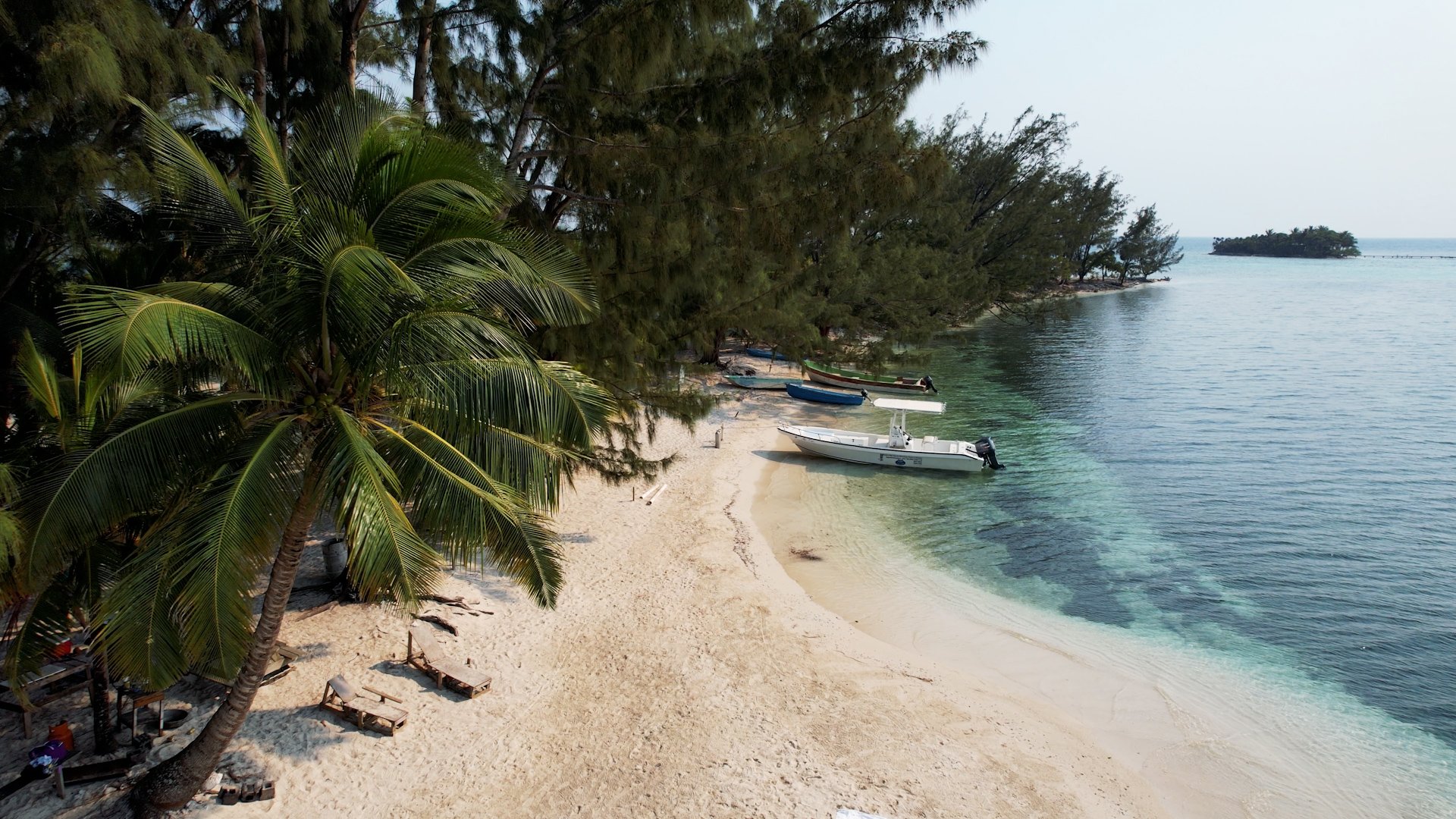 Tropical beach with white sand, several boats anchored near the shore, lush green trees including palm trees, and calm turquoise water.