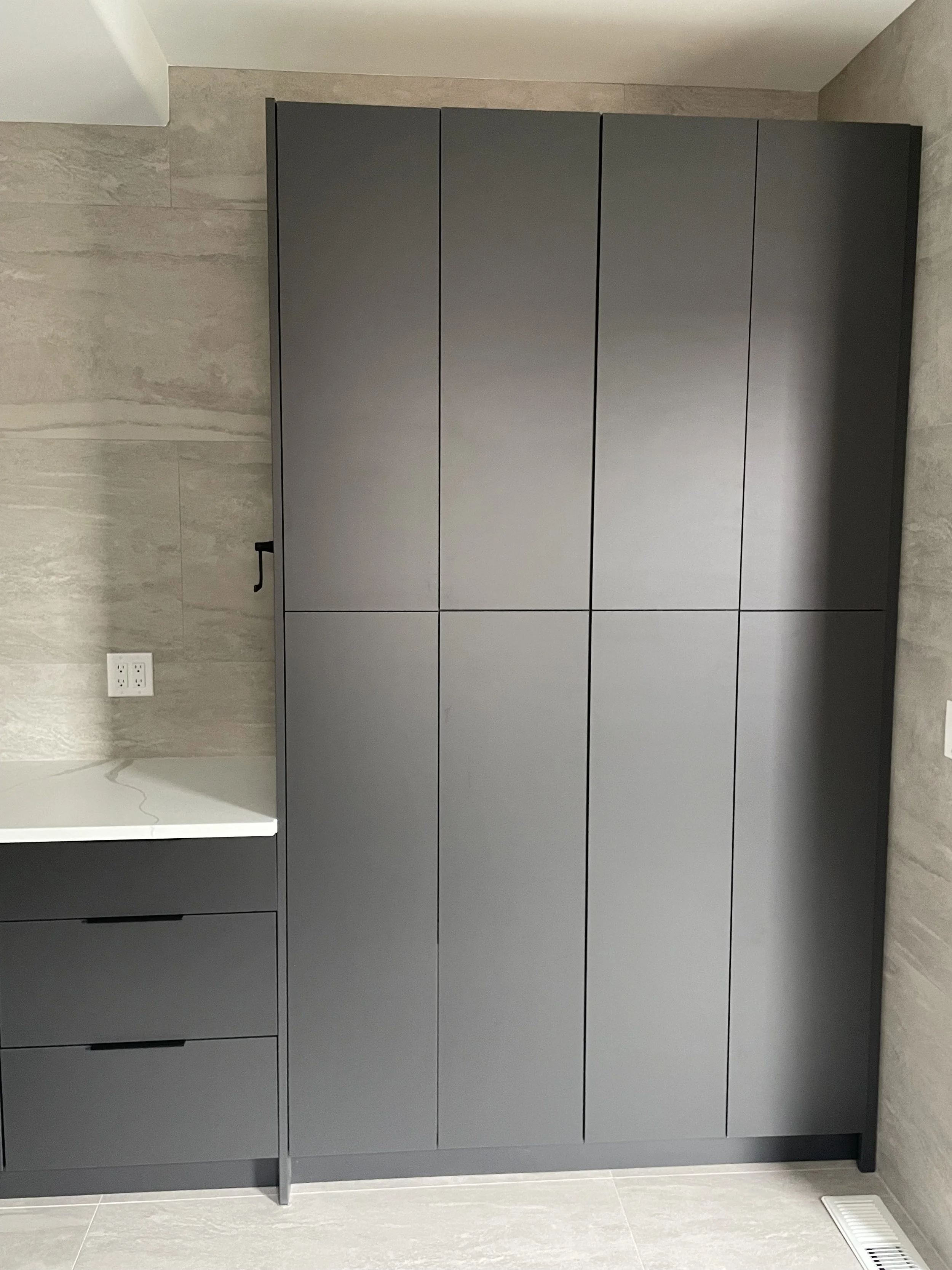 A tall, dark gray cabinet with six vertical doors is placed against a wall in a modern room. Next to it is a smaller cabinet with three drawers and a white countertop. The room has beige, textured wall tiles and a light-colored tiled floor.