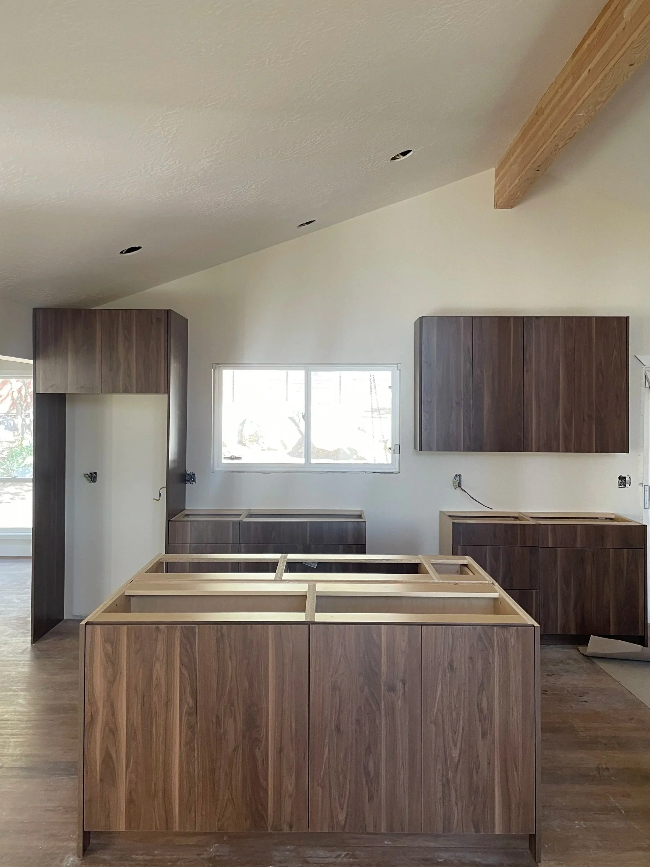 Kitchen under construction with wooden cabinets and island in center, partially assembled, with upper cabinets on wall and a window above.