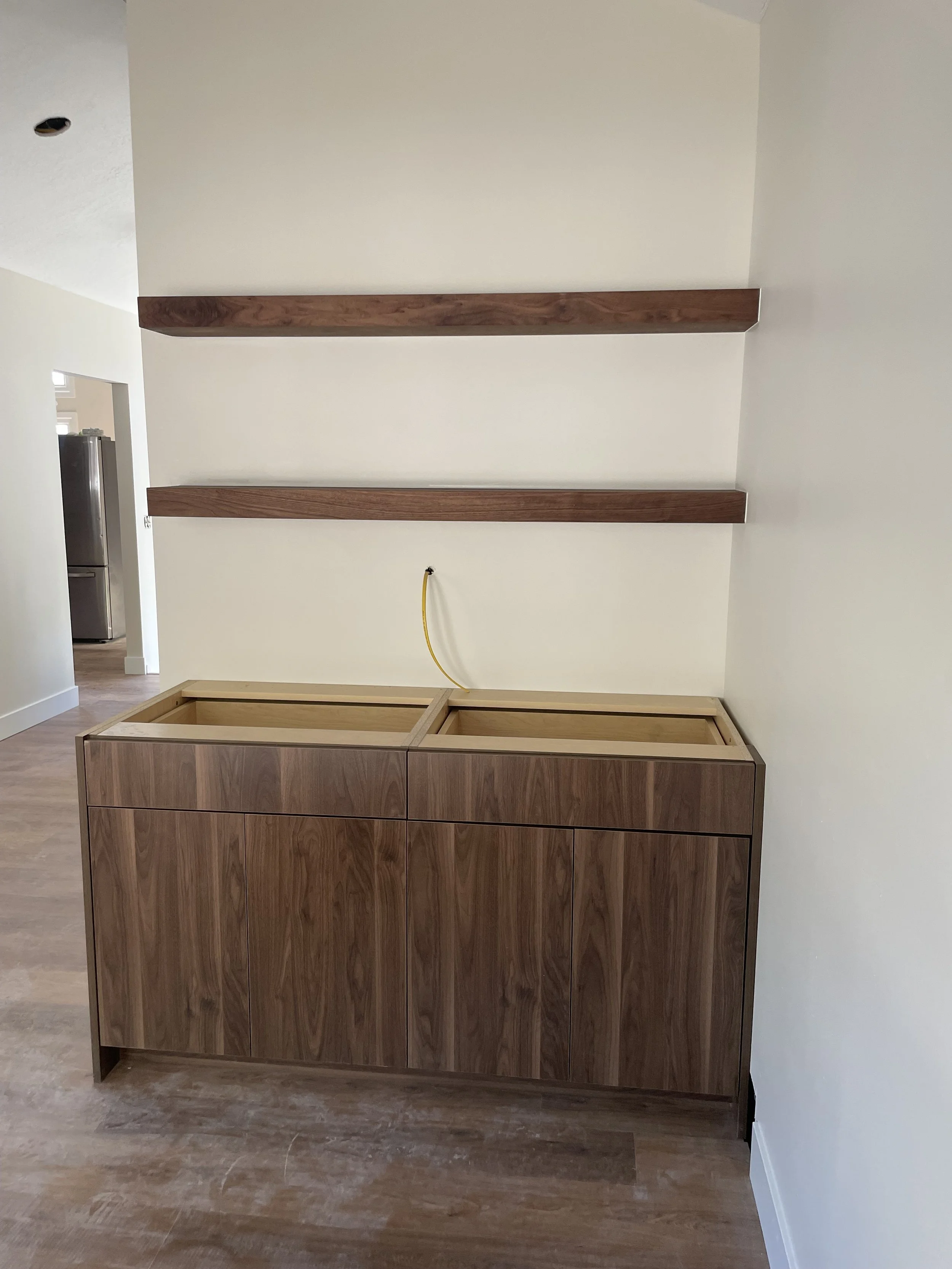 Empty wooden cabinet with two compartments, two wooden shelves mounted on the wall above, and a yellow hose connected to a water pipe.