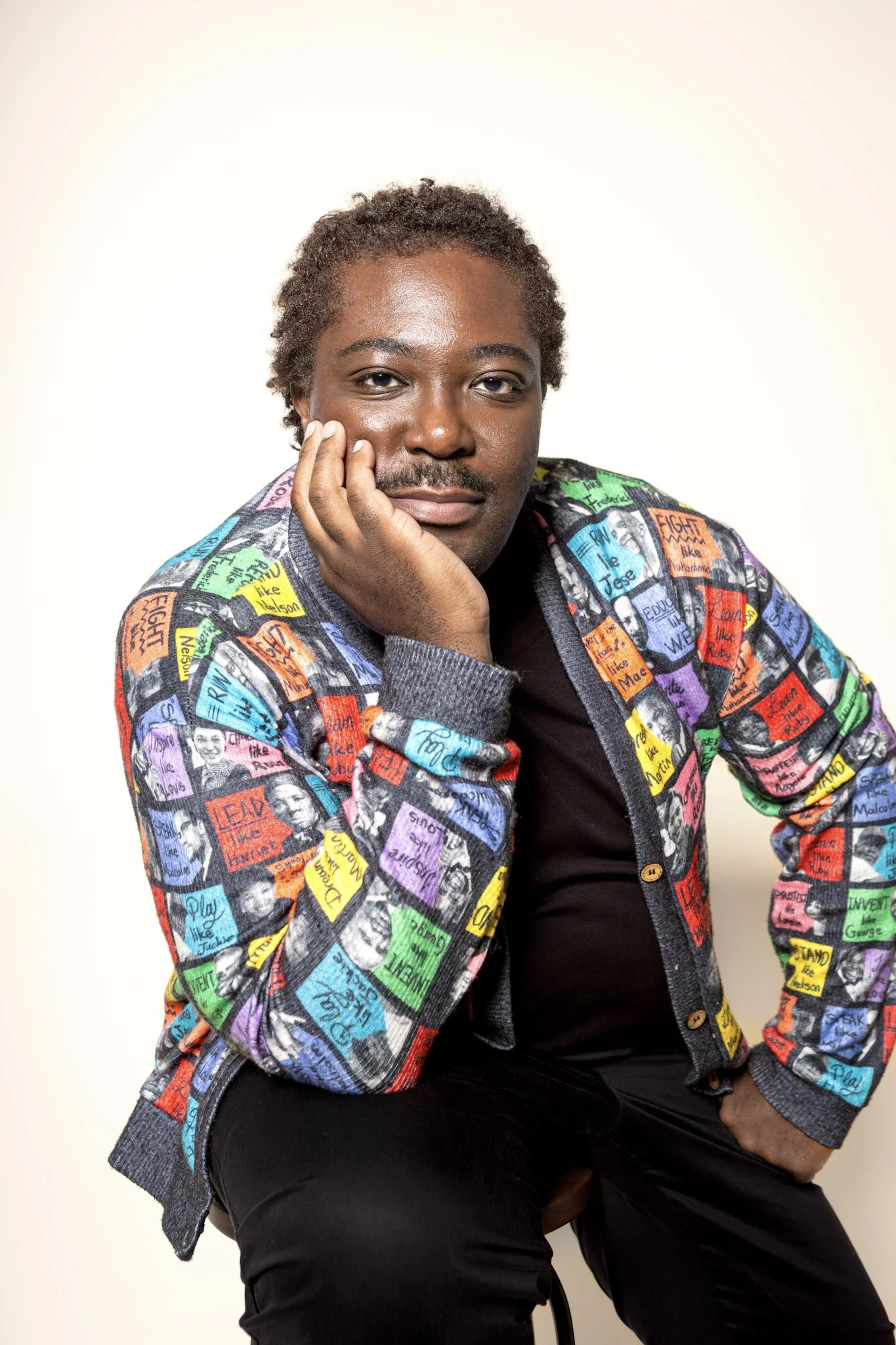 Portrait of Calvin Eaton seated against a light background, wearing a colorful patterned sweater and resting his face on his hand
