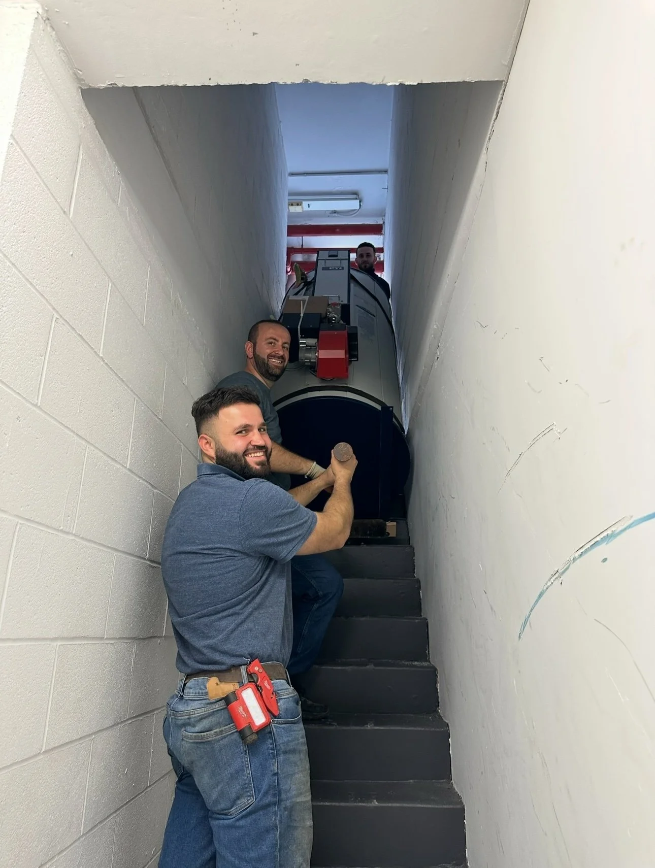 Three men are moving a large piece of equipment up a narrow staircase. They are smiling and working together.