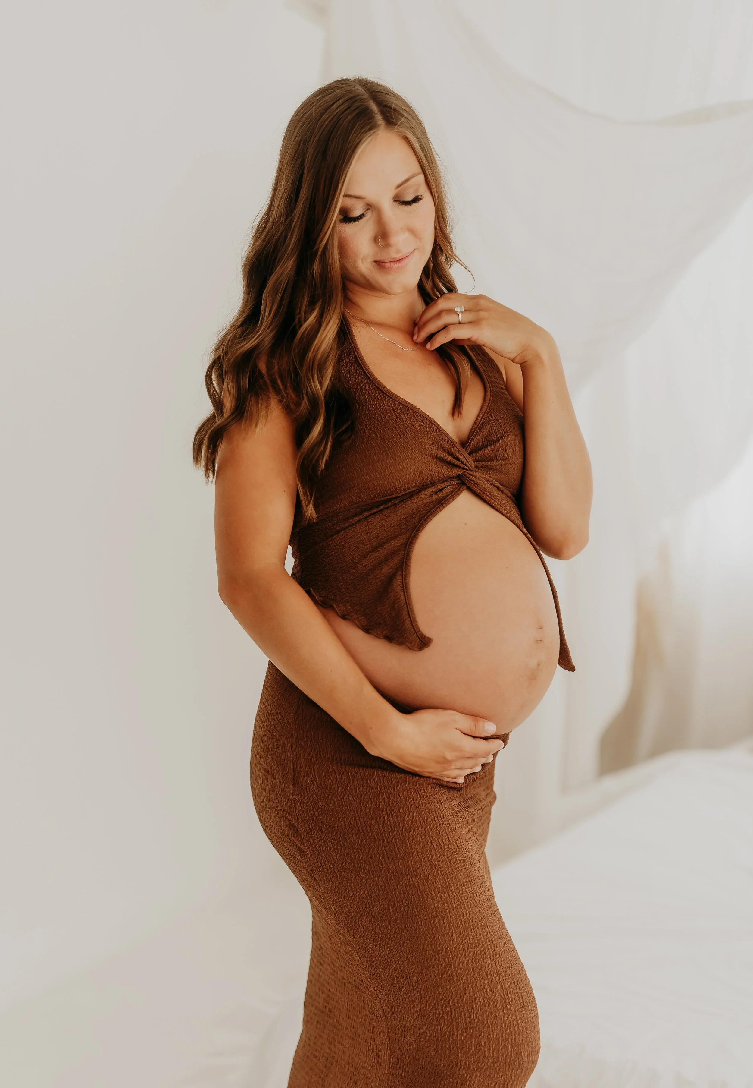 Pregnant woman in an off-shoulder dress leaning against a wall, hand on her belly, looking to the side.