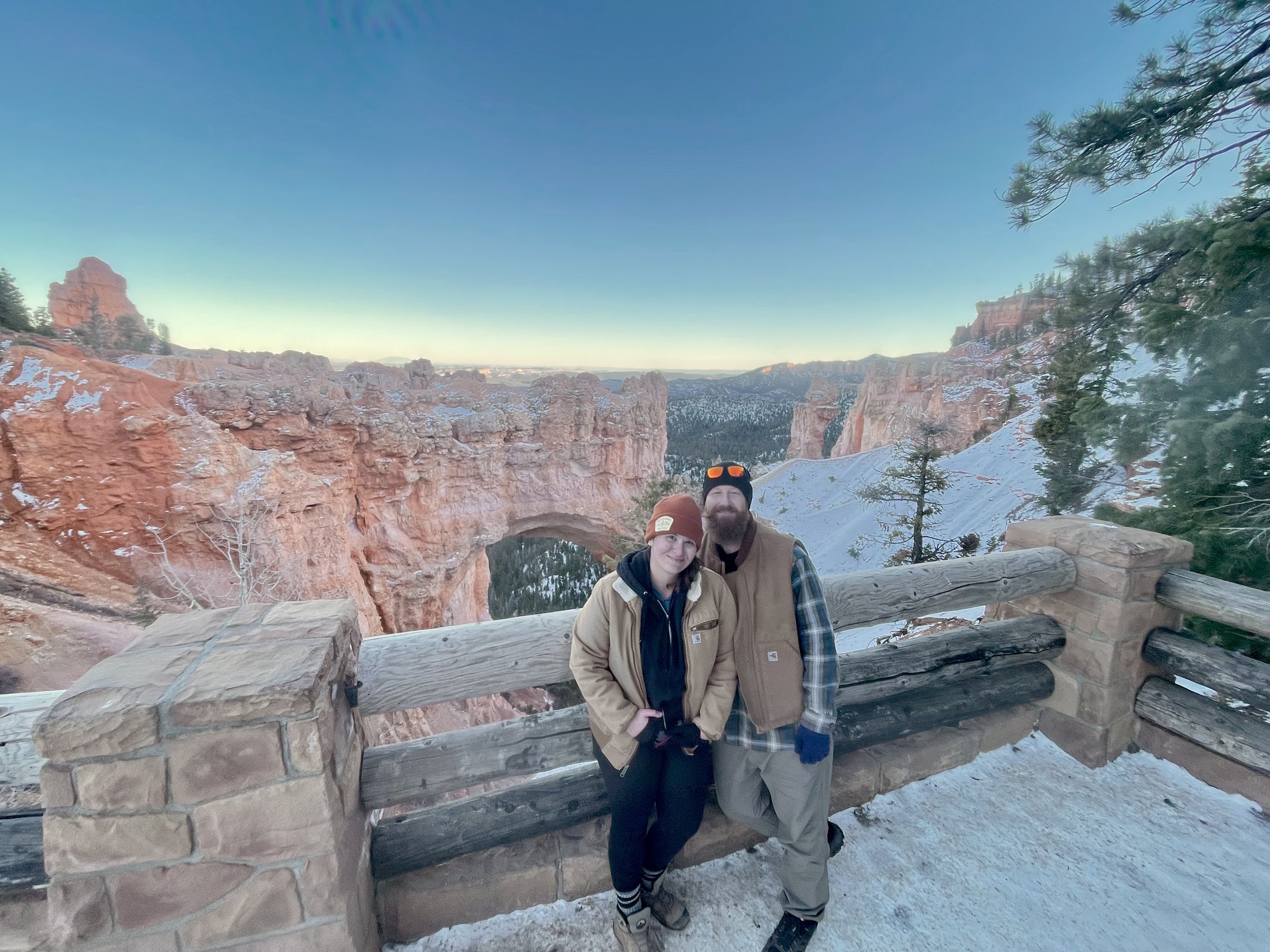A smiling couple posing by a wooden and stone railing at a scenic overlook in a snowy canyon with red rock formations and evergreen trees during sunset.