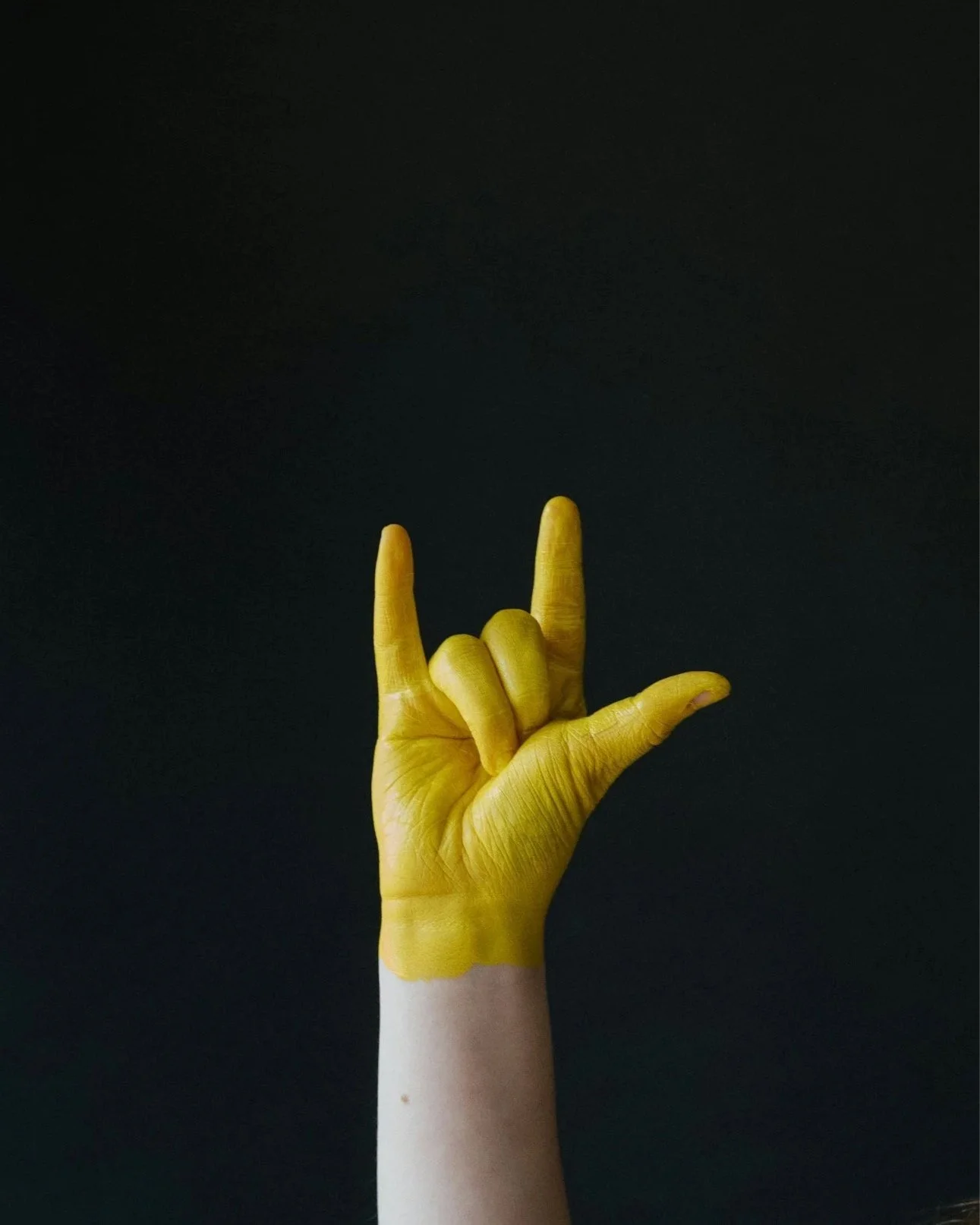 Person's hand painted yellow with the thumb, index, and pinky fingers extended in a I love you American Sign Language gesture against a black background
