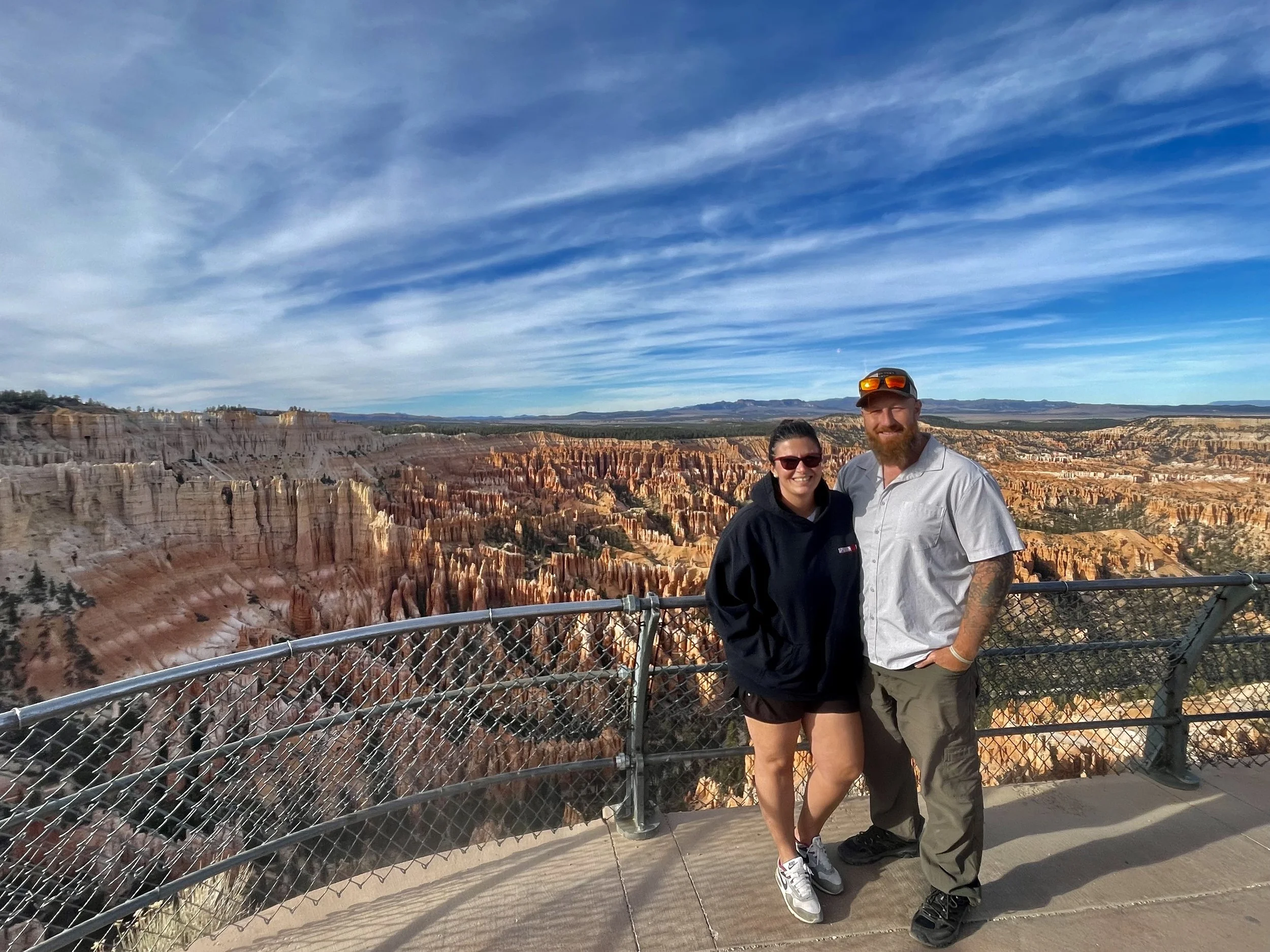 A smiling woman and man standing together at a scenic overlook with a canyon and colorful rock formations in the background, under a blue sky with wispy clouds.